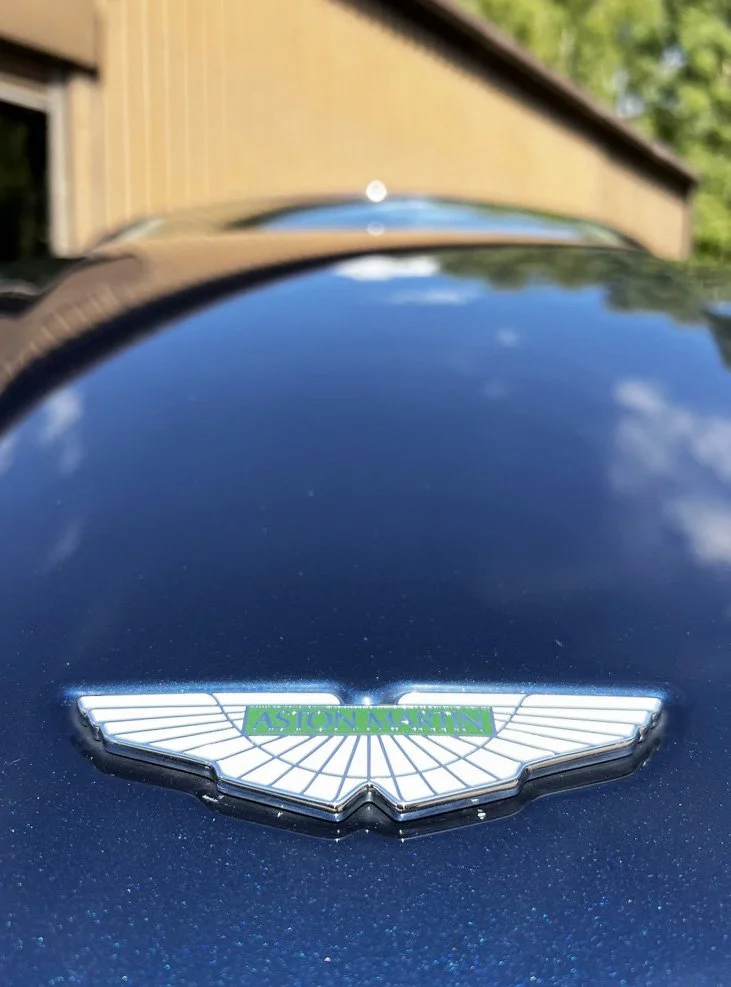 Close-up of an Aston Martin emblem on the front of a dark blue car, with building and trees in the background