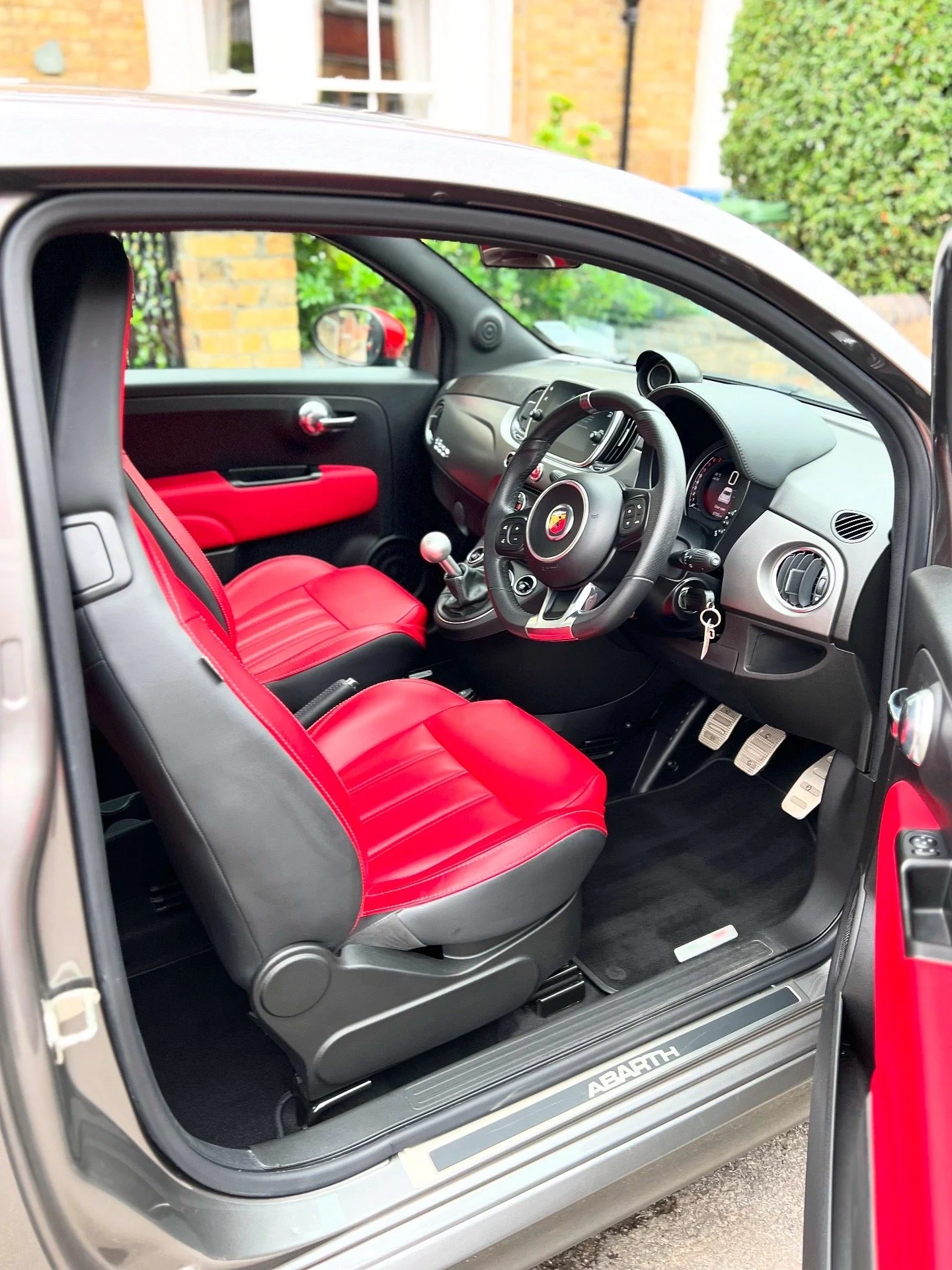 Interior of a compact car with red and black seats, dashboard, steering wheel, and gear shift, parked outside near a house with greenery.