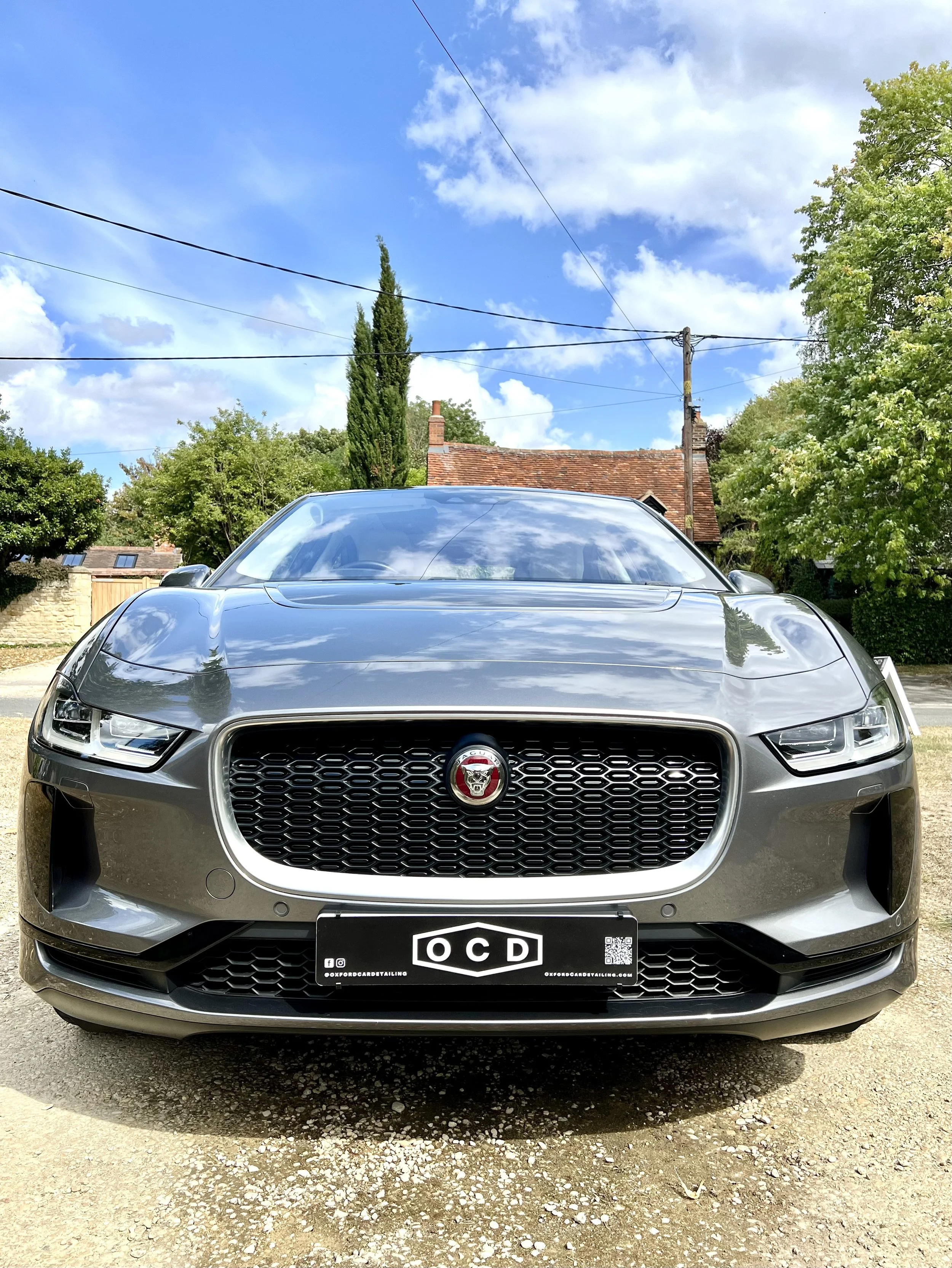 Front view of a Jaguar car with a black OCD license plate, parked outdoors with trees and a house in the background, under a partly cloudy sky after full valet and detailing.