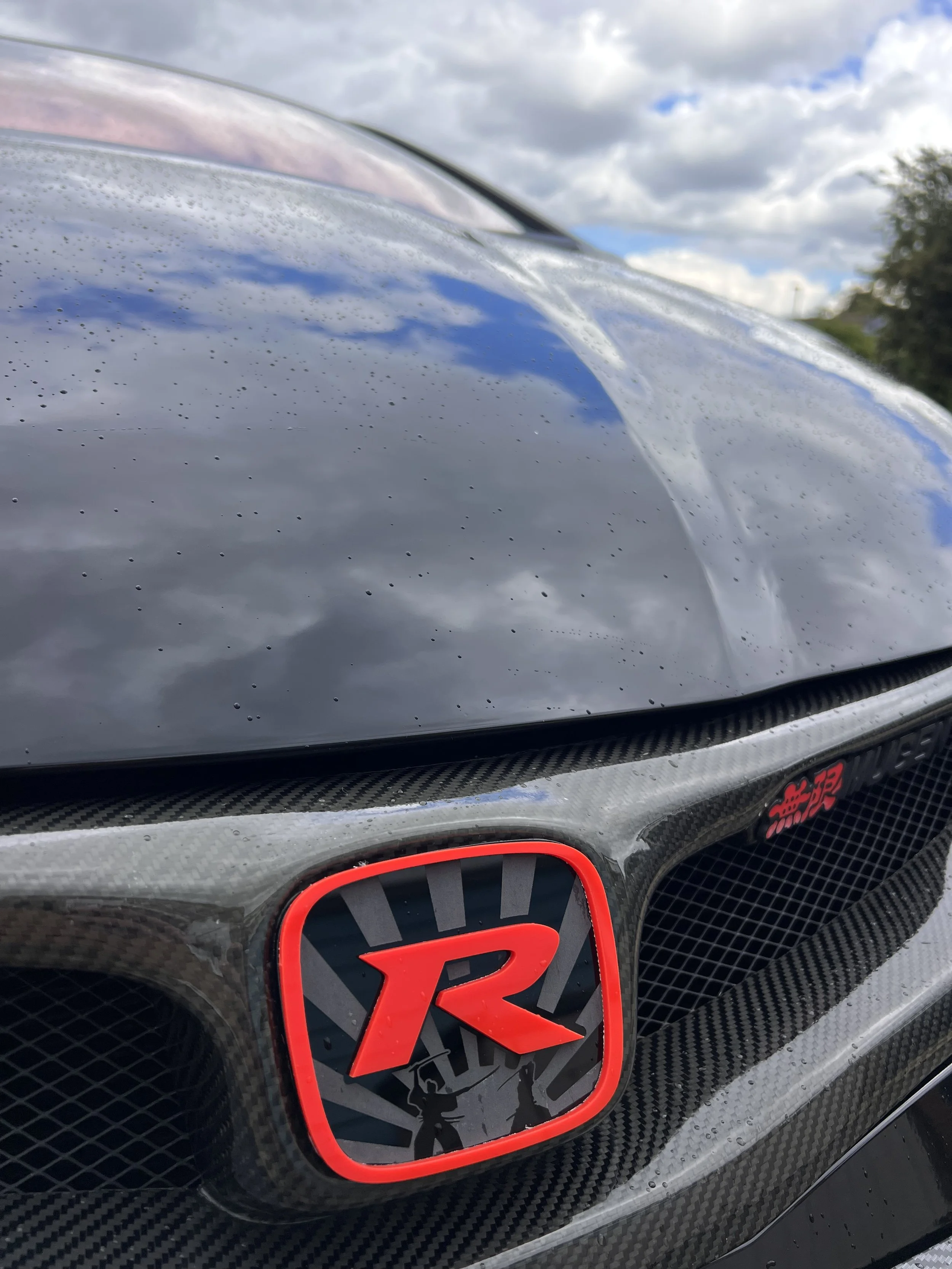 Close-up image of the front of a black sports car with a red trademark emblem and a carbon fiber grille. Droplets of water are visible on the car's surface, indicating it has been recently washed or rained on. The sky with clouds can be seen reflecte