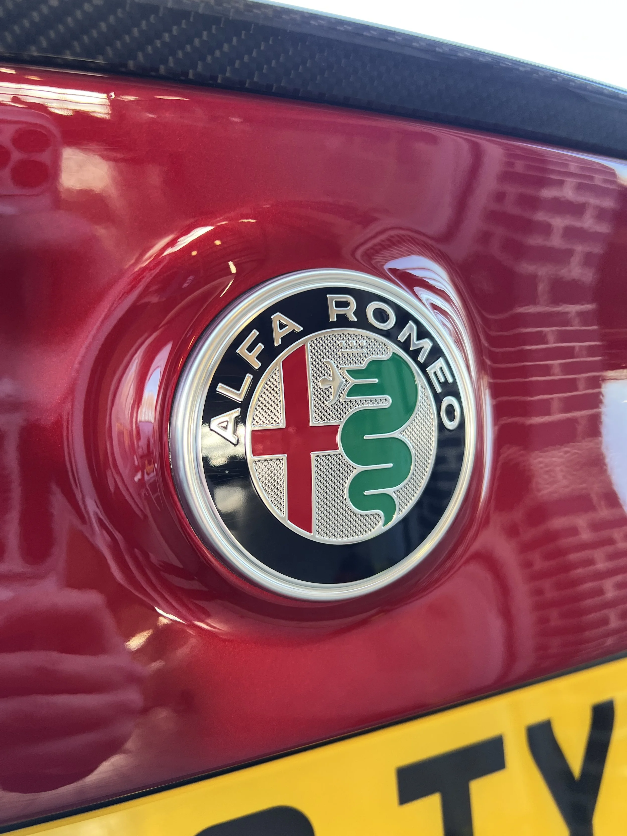 Close-up of an Alfa Romeo emblem on a red car with a partial yellow license plate visible at the bottom.