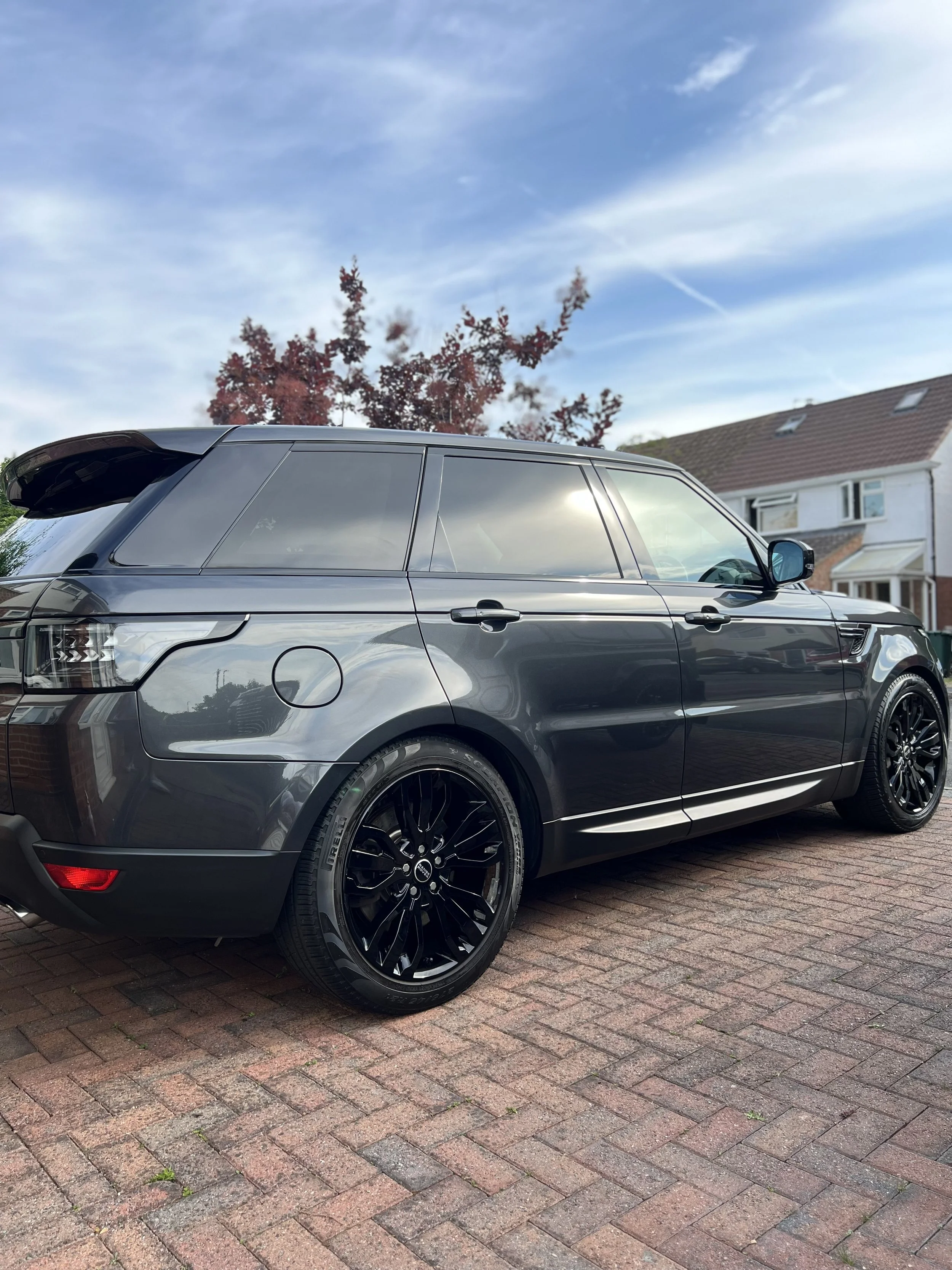 A dark gray Range Rover SUV parked on a brick driveway with houses and a blue sky in the background.