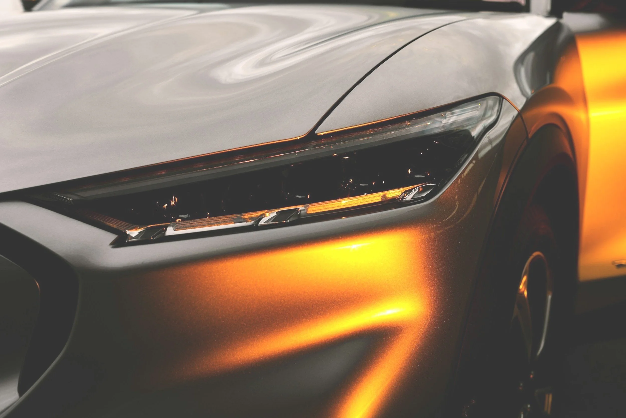 Close-up of the front left side of a silver and orange sports car, showing the headlight and part of the hood and wheel, with a sleek and modern design.
