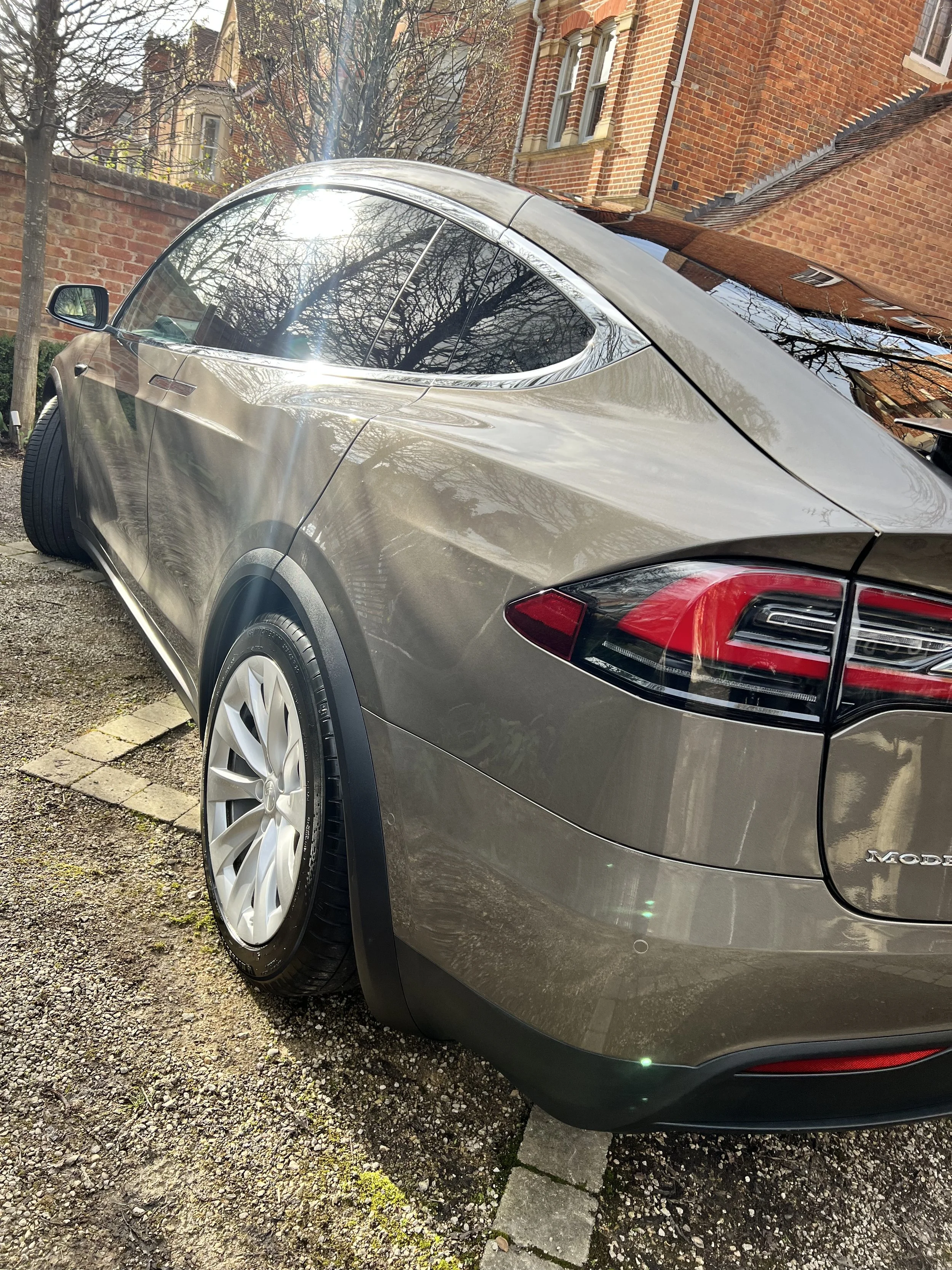 A silver Tesla Model Y parked on a gravel driveway in front of brick buildings and trees with bare branches.