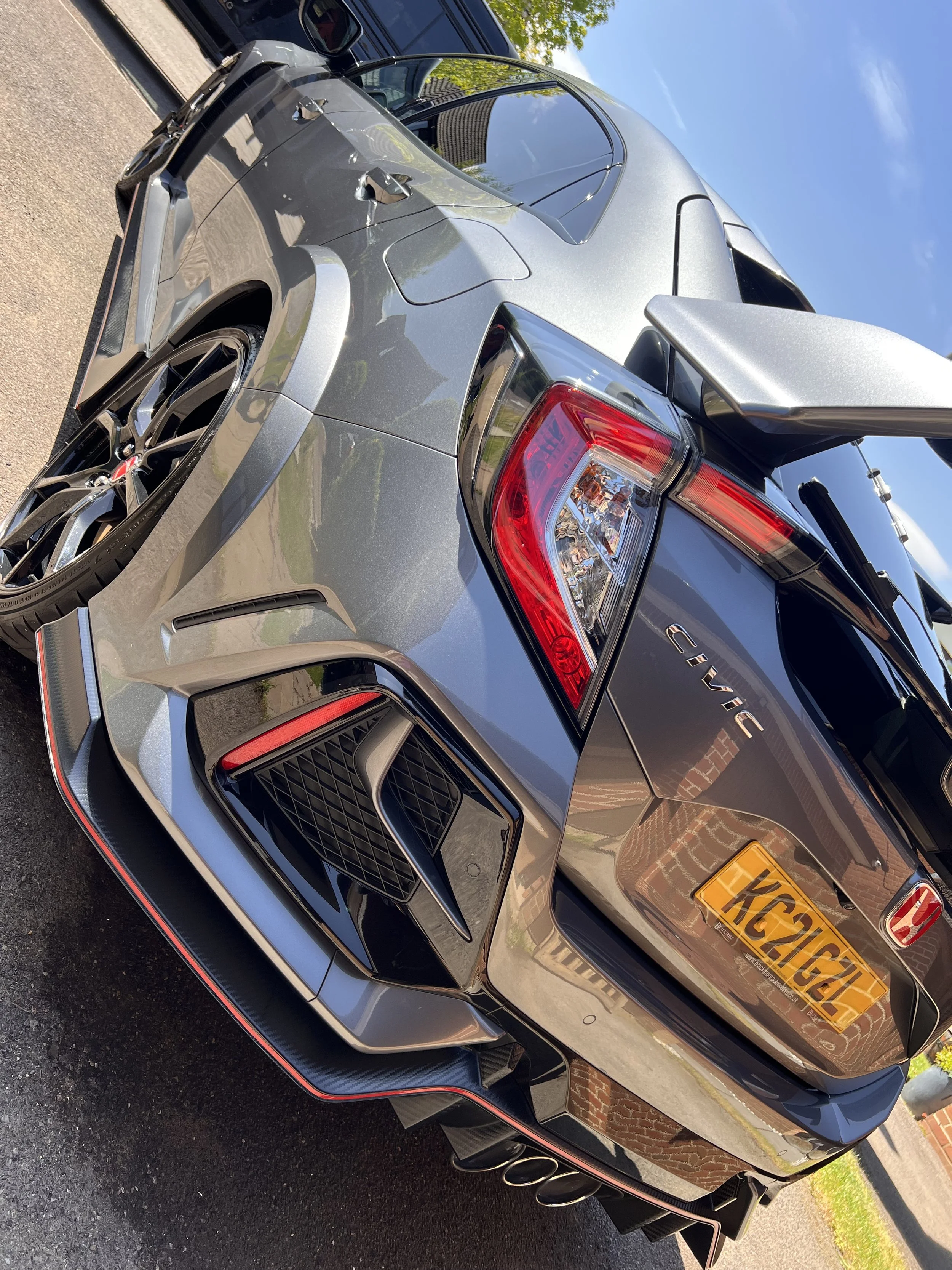A silver Mercedes-AMG GT parked outdoors next to a brick building with a yellow license plate reading 'H624GAA'. The car has a visible AMG badge, black and silver wheels, black front grille, and a rear wing spoiler.
