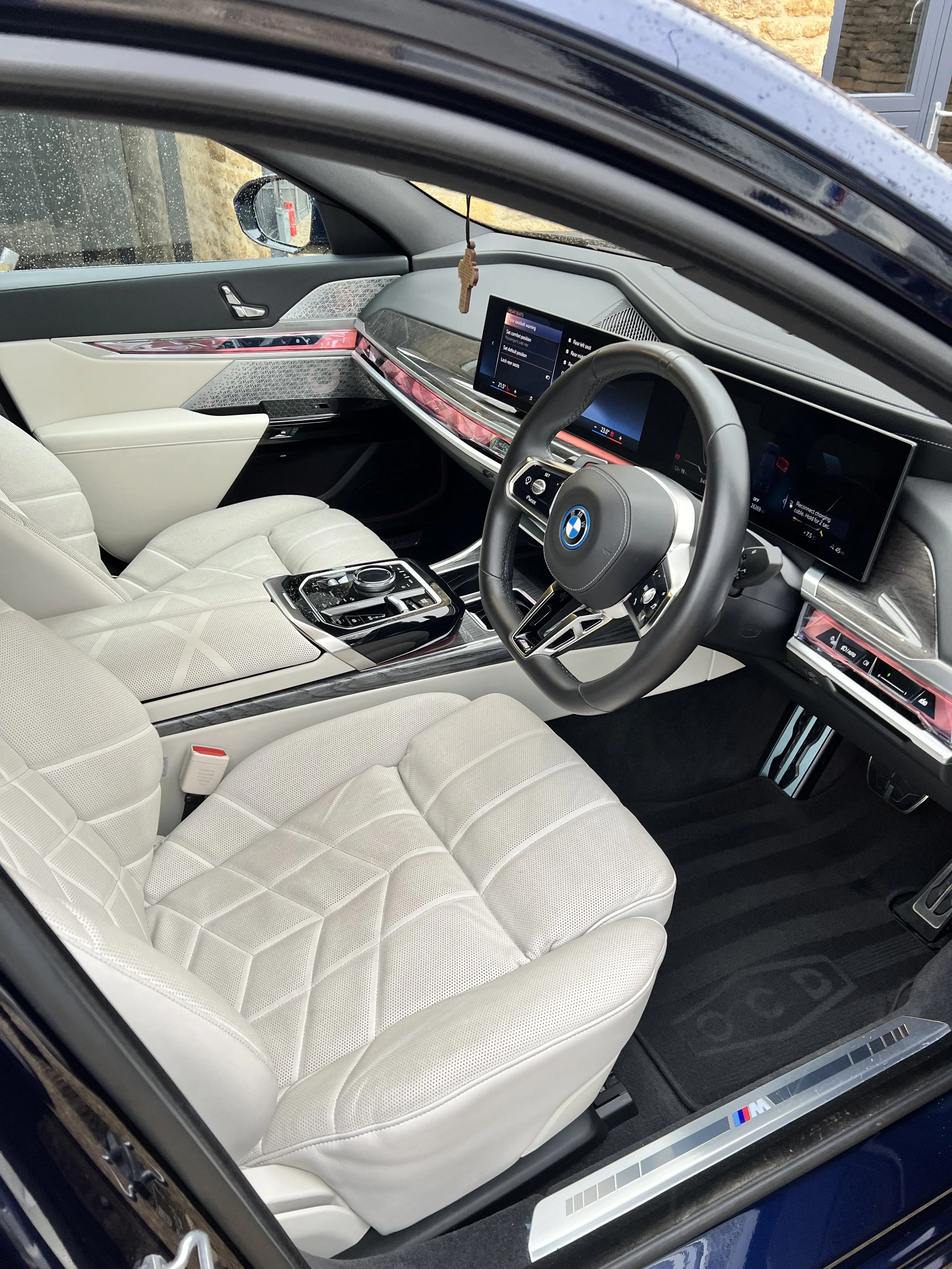 Interior of a BMW car with beige seats, a digital dashboard, and steering wheel, with a dash-mounted control pad, and rain droplets on the window after full valet and detail and ceramic coating.