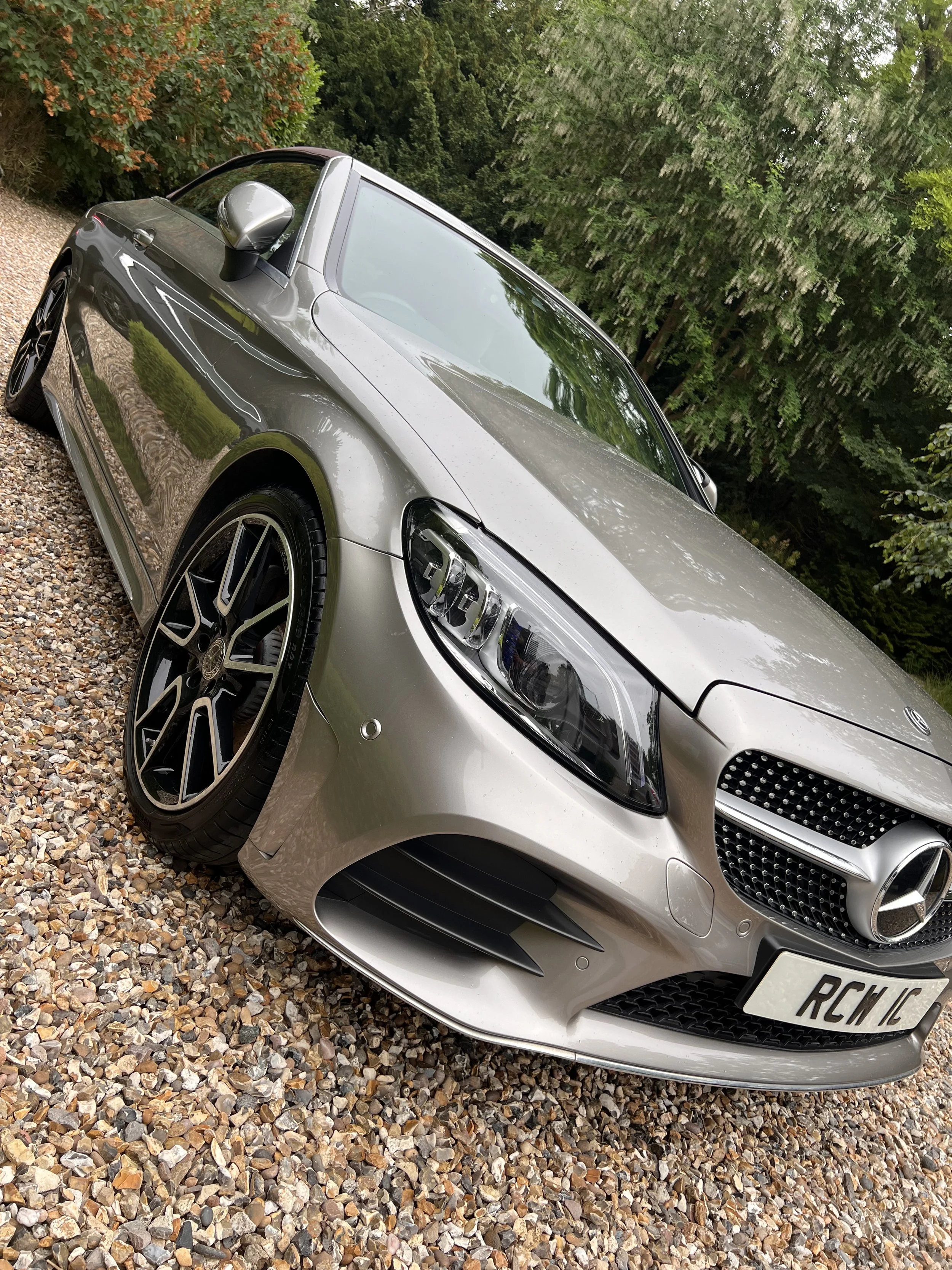 A silver Mercedes-Benz sedan parked on a gravel driveway with greenery and trees in the background.