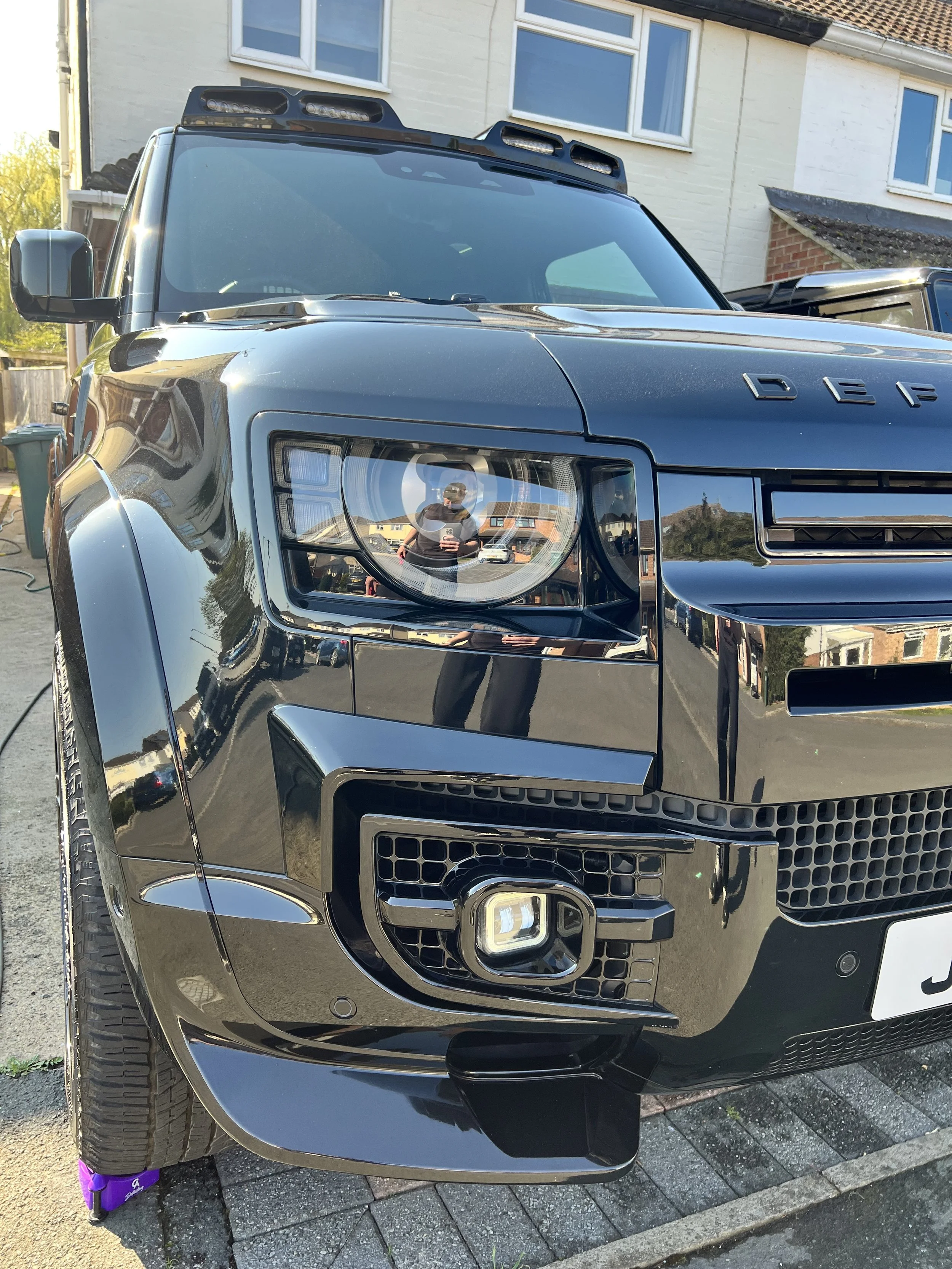 Close-up of a black Range Rover SUV parked on a paved driveway in front of a residential house with white brick walls and large windows. The reflection of the person taking the photo, wearing a gray shirt and sunglasses, is visible in the car's headl