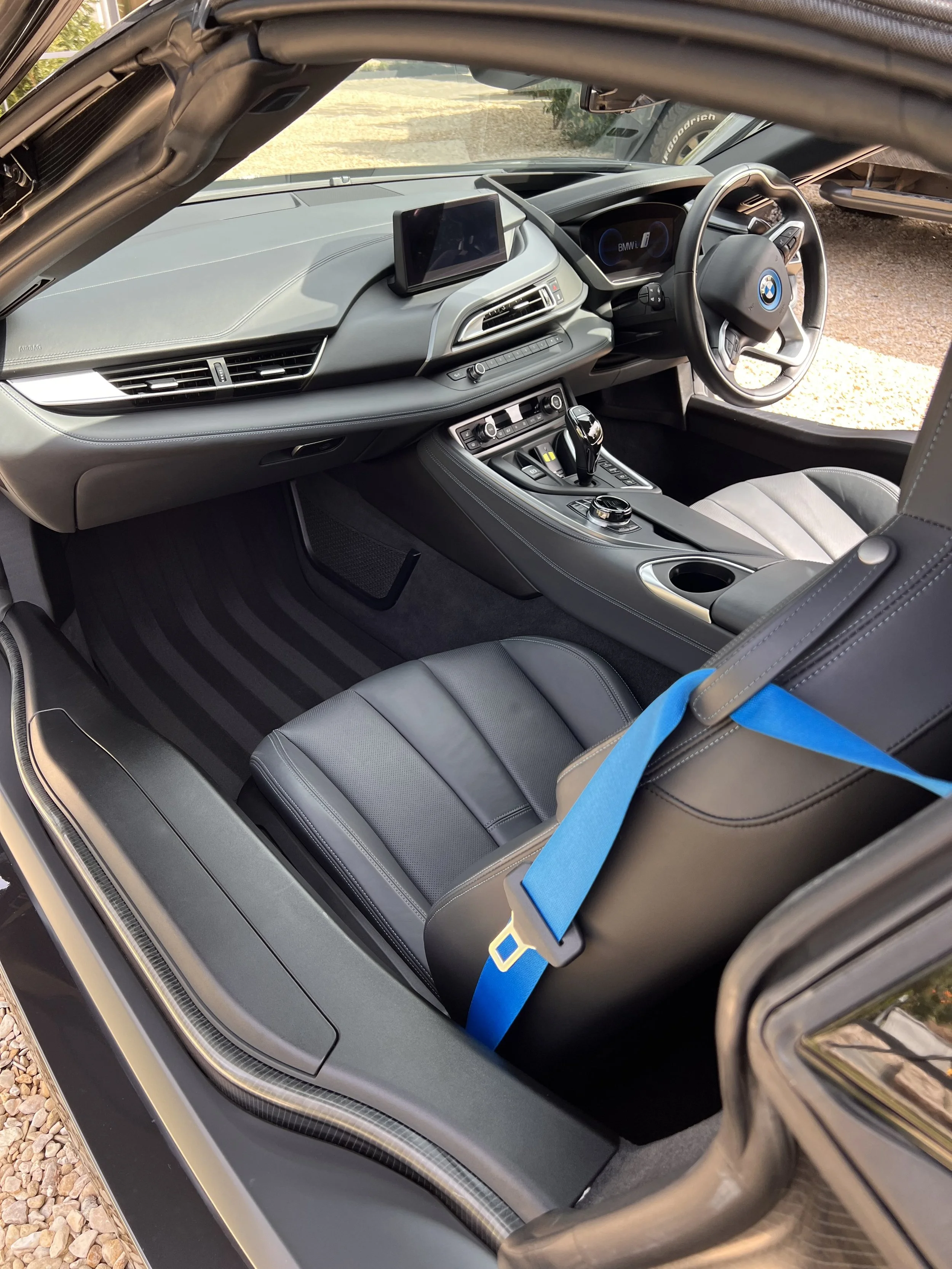 Interior of a BMW car showing the dashboard, steering wheel, gear shifter, and black leather seats, with a blue seatbelt.