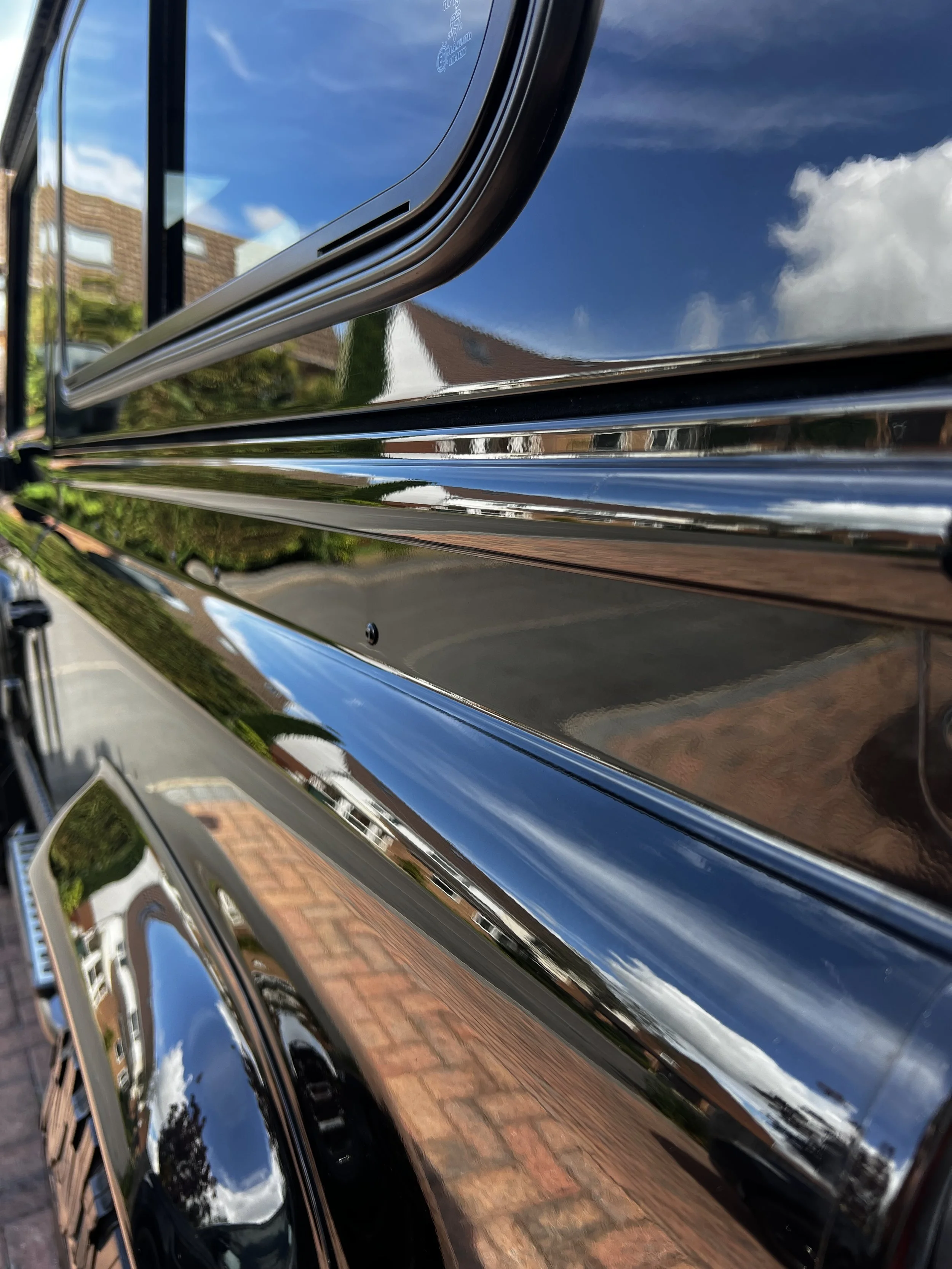 Close-up of a black car's glossy exterior with reflections of clouds, blue sky, and neighborhood houses.