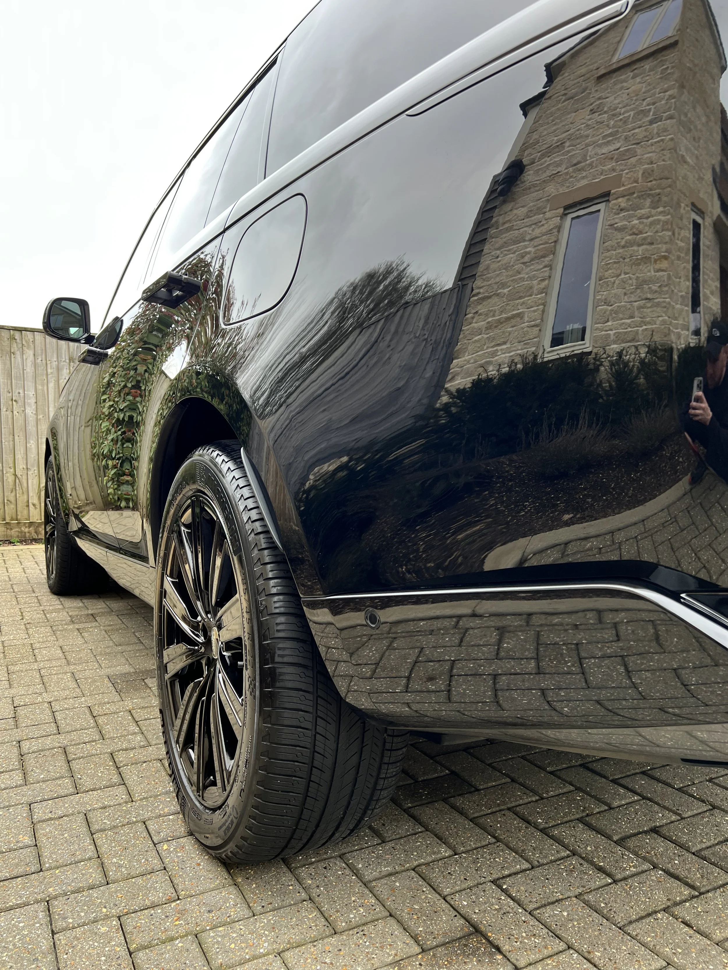 Close-up of a black car with shiny, reflective surface parked on a brick driveway, showing only part of the side and wheel, with a stone building and cloudy sky reflected on its surface.