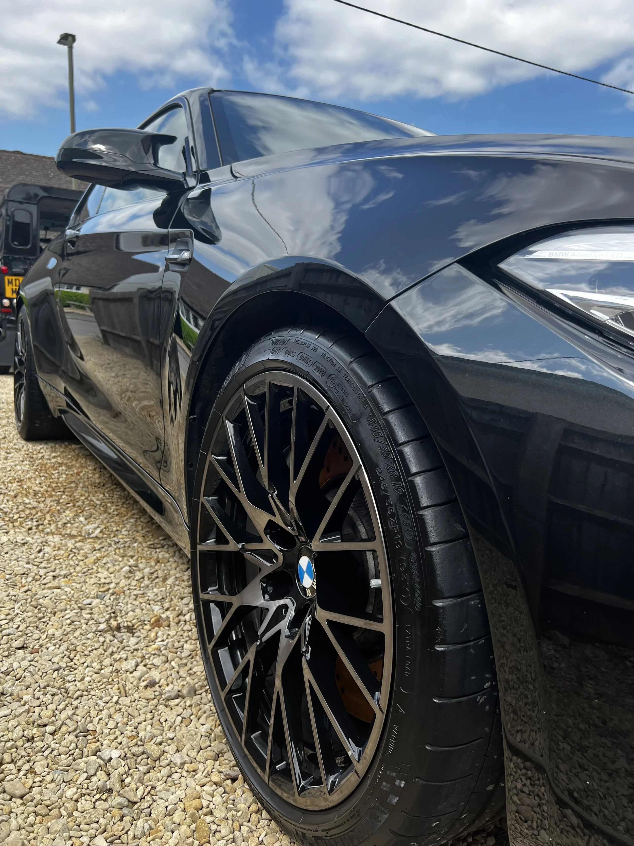 Close-up of a black BMW car with shiny finish, showing the front wheel, part of the hood, and side mirror, with a partly cloudy sky reflection.