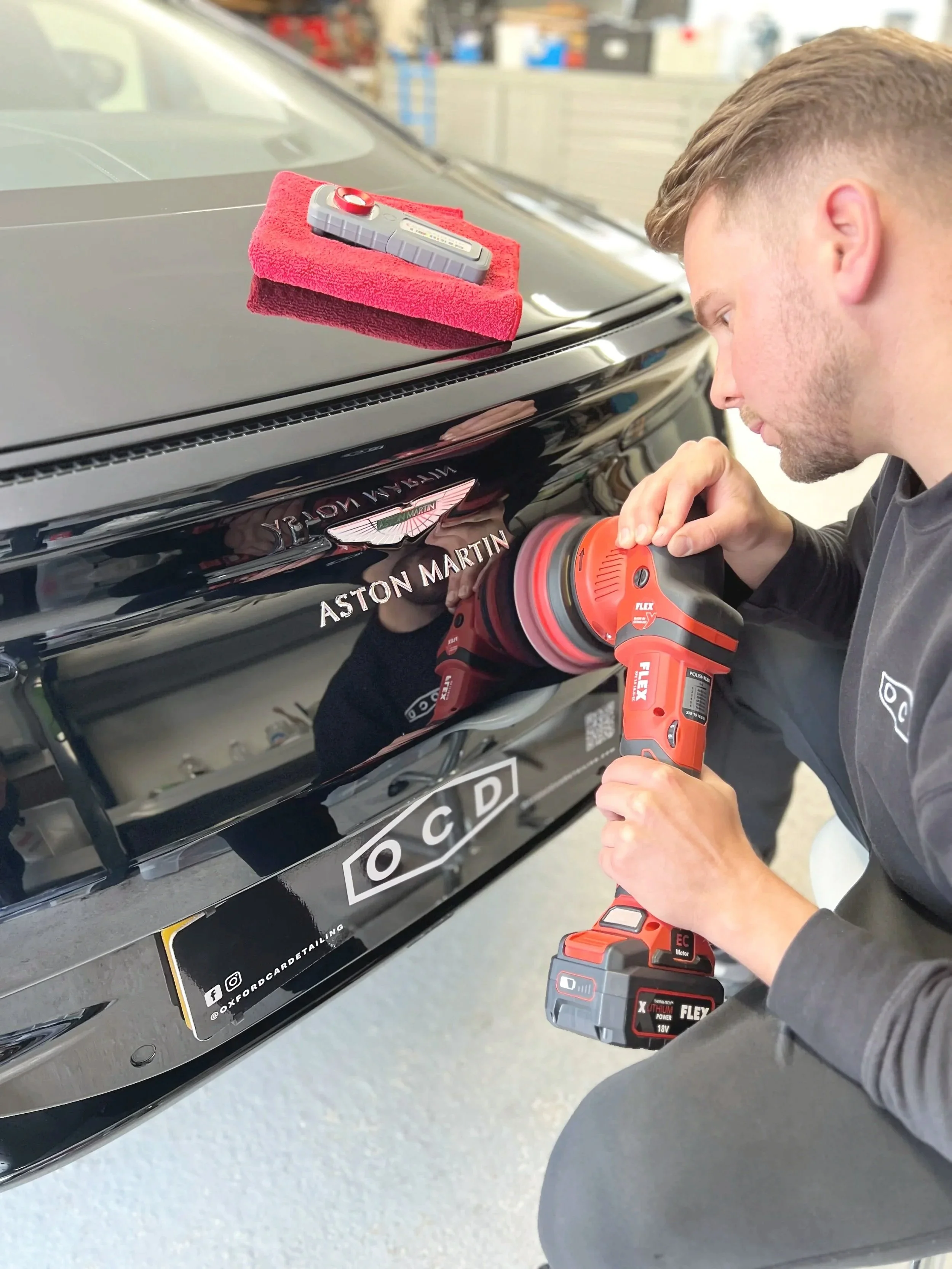 Paint correction via machine polishing a black Aston Martin car with a red and black cordless power buffer in a garage.