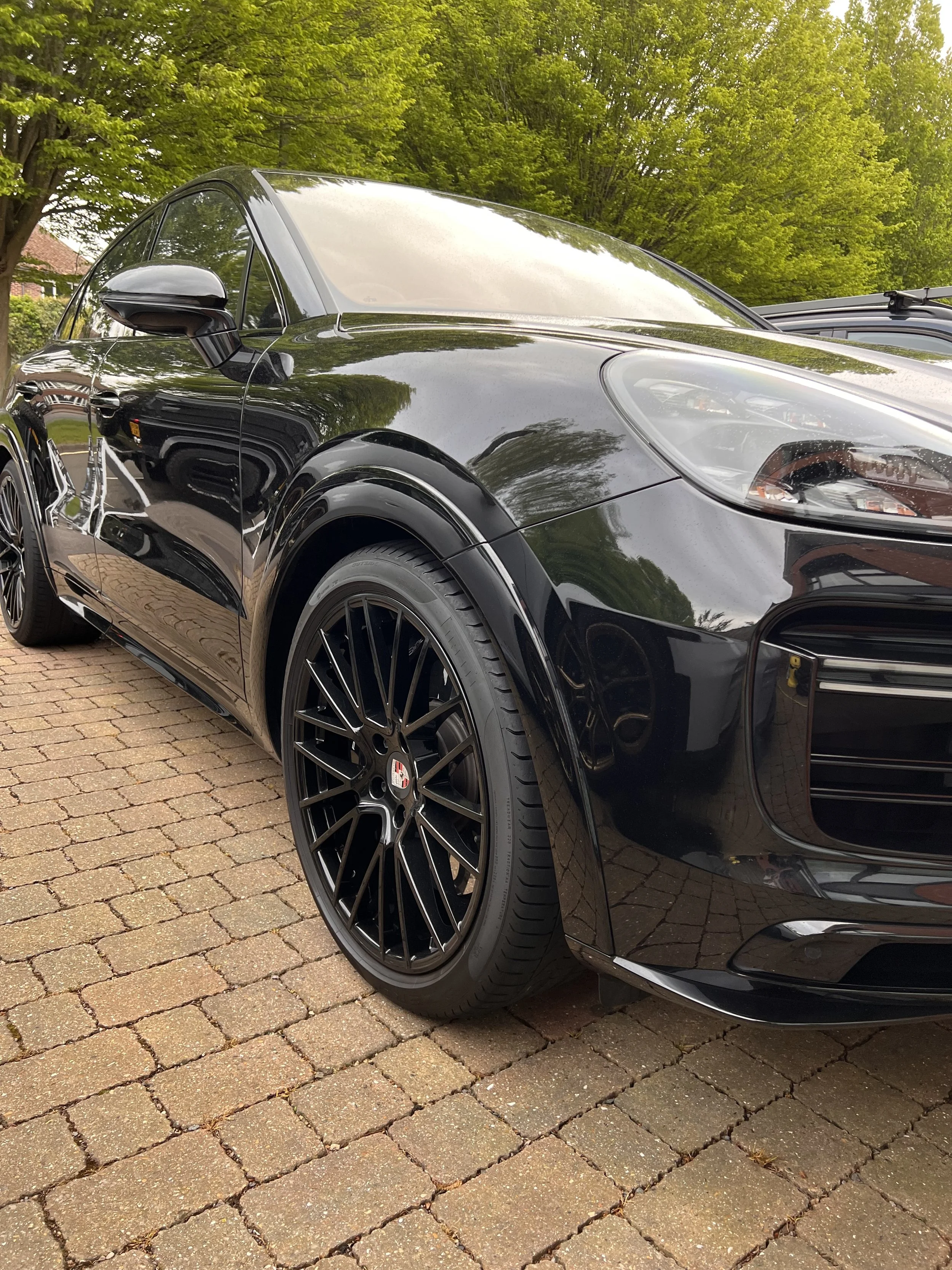 A black sports car with black wheels parked on brick pavement, with green trees in the background.