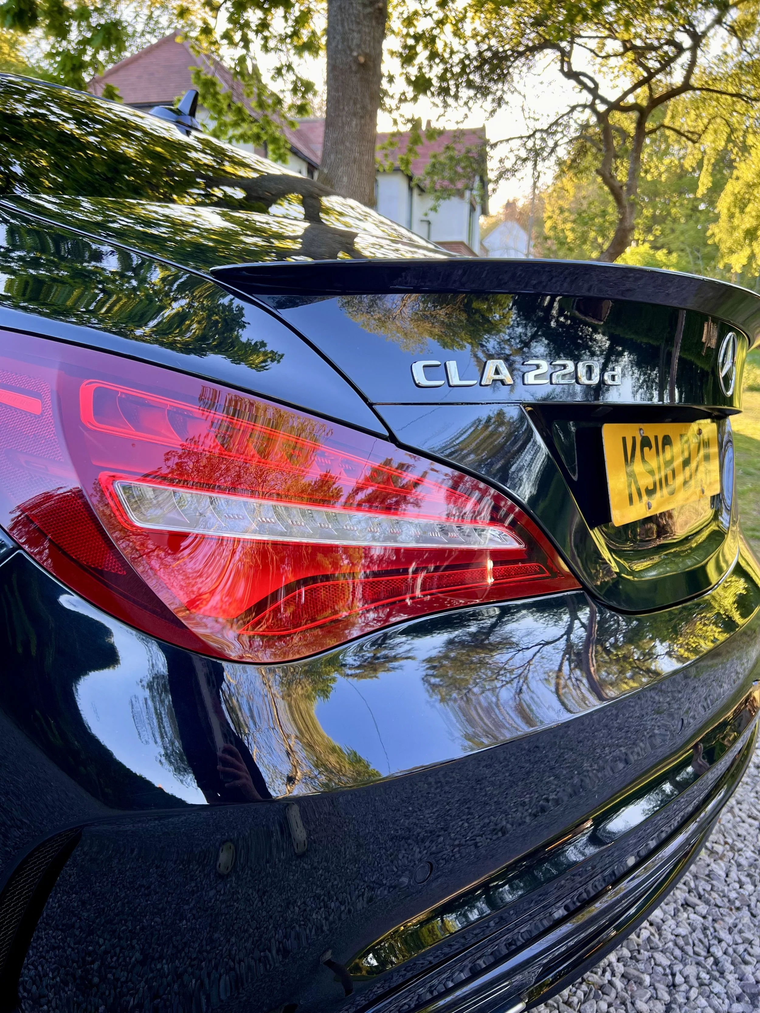 Close-up of the back of a black Mercedes-Benz CLA 220d with reflections of trees and houses on its glossy surface, showing the rear lights, model badge, and license plate.