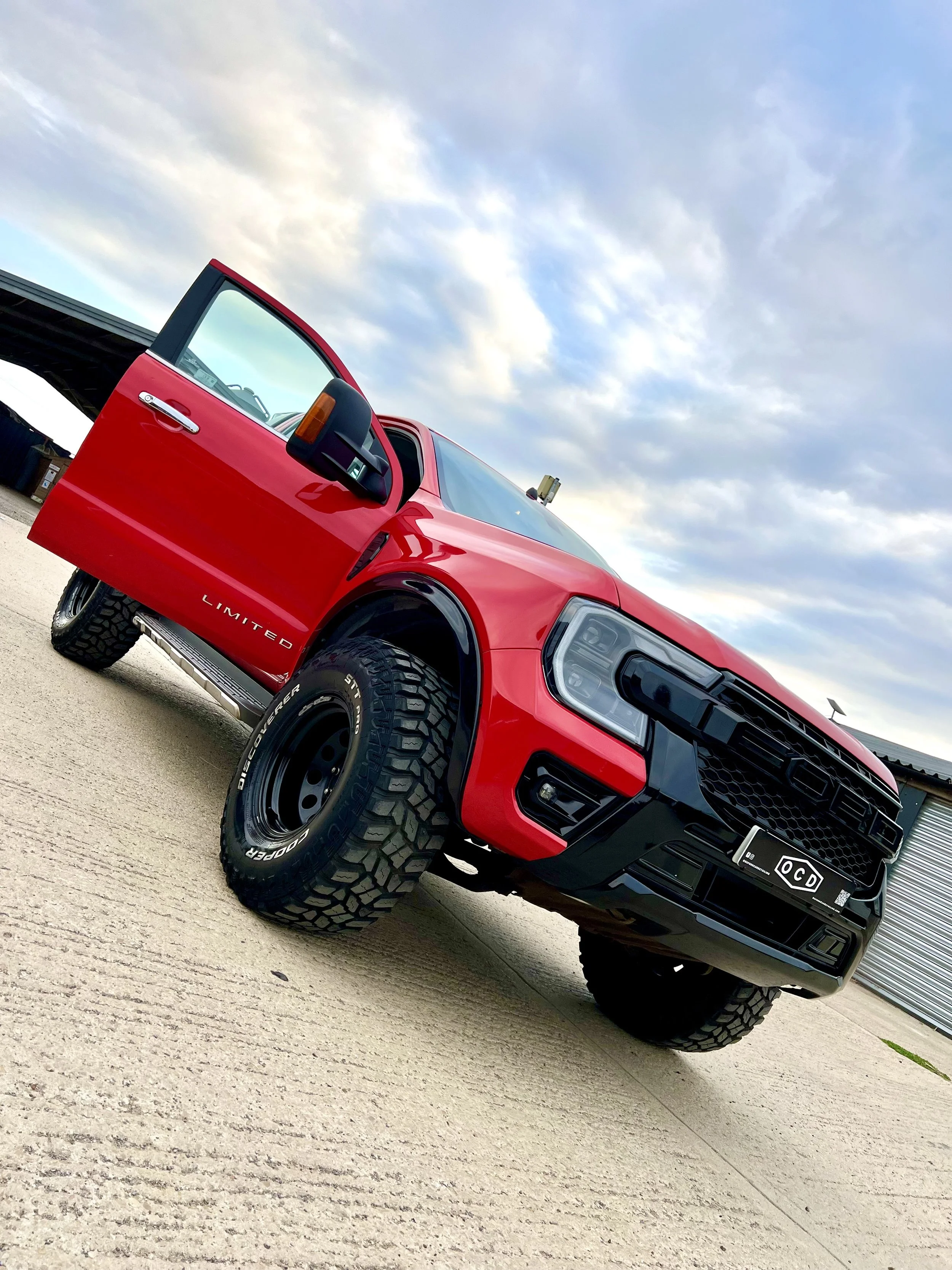 Red Ford pickup truck with open driver's side door parked on concrete surface under cloudy sky.