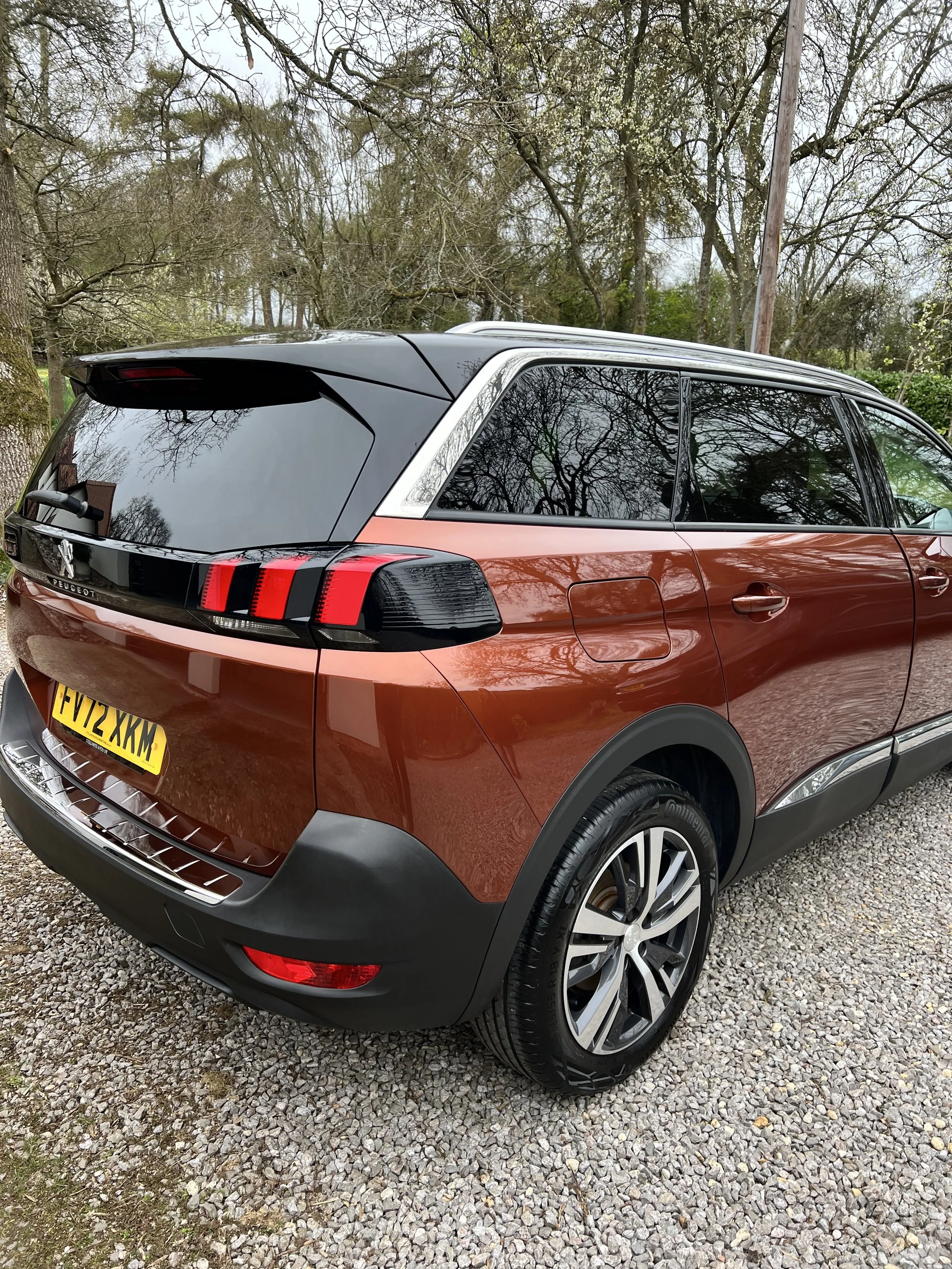 A red and black Peugeot SUV parked on a gravel driveway surrounded by leafless trees.