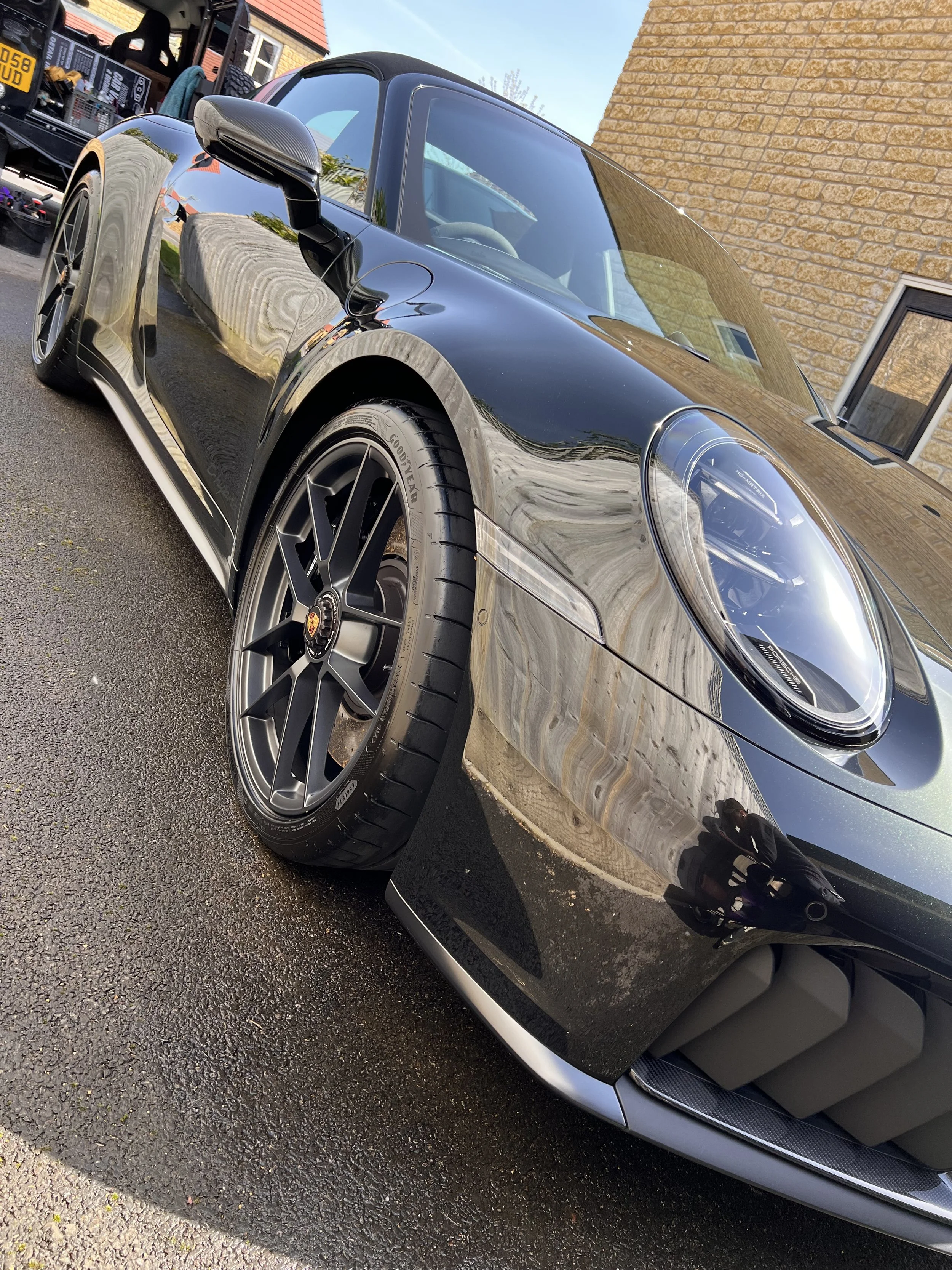 Black Porsche sports car parked on a driveway outside a brick house with a sloped roof, simple window, and glass door, reflecting the sky and nearby surroundings.