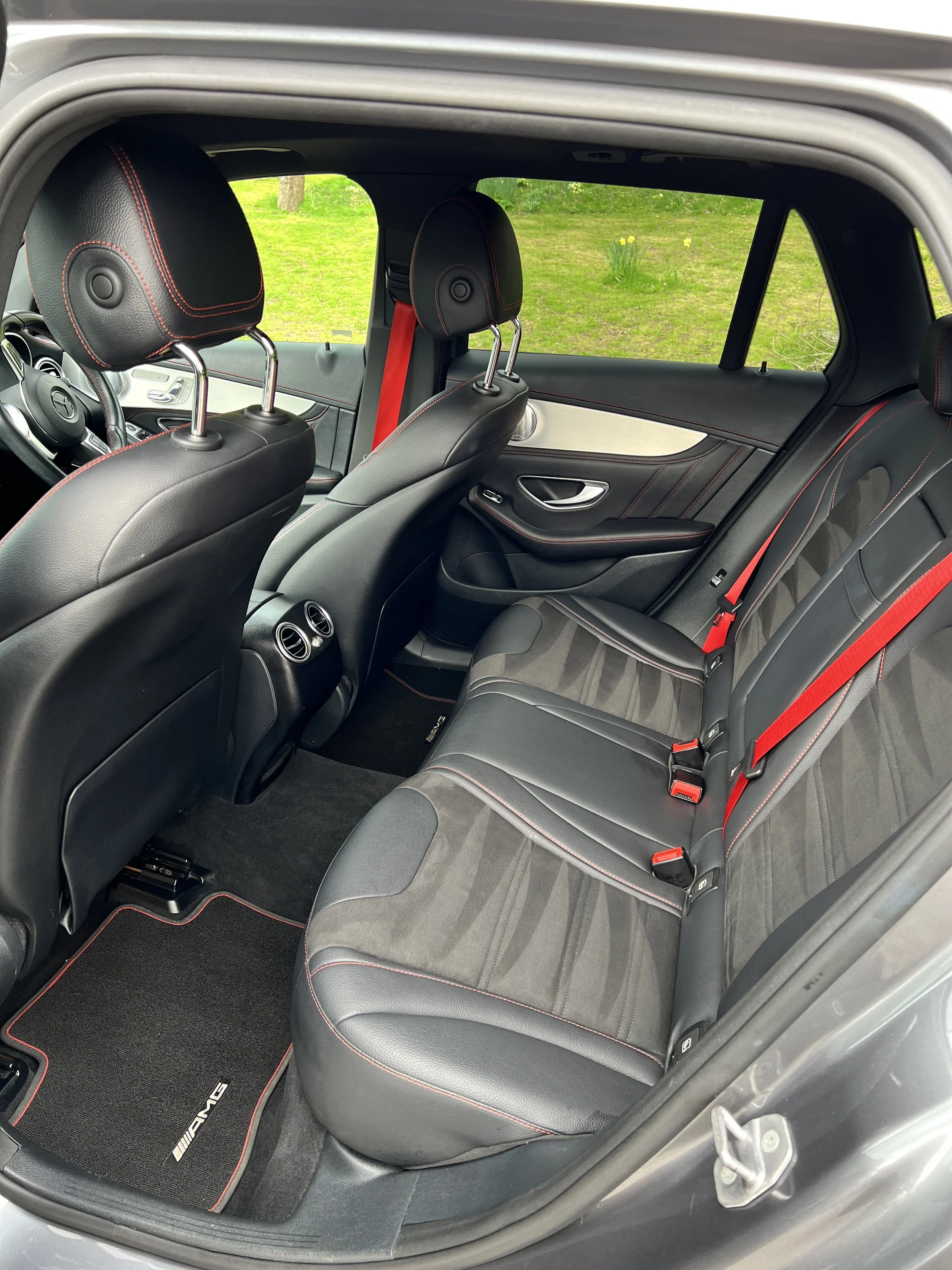 The interior of a Mercedes-Benz car showing black leather seats with red stitching, headrests, a rear bench seat with red seat belts, and a floor mat with Mercedes-AMG branding.