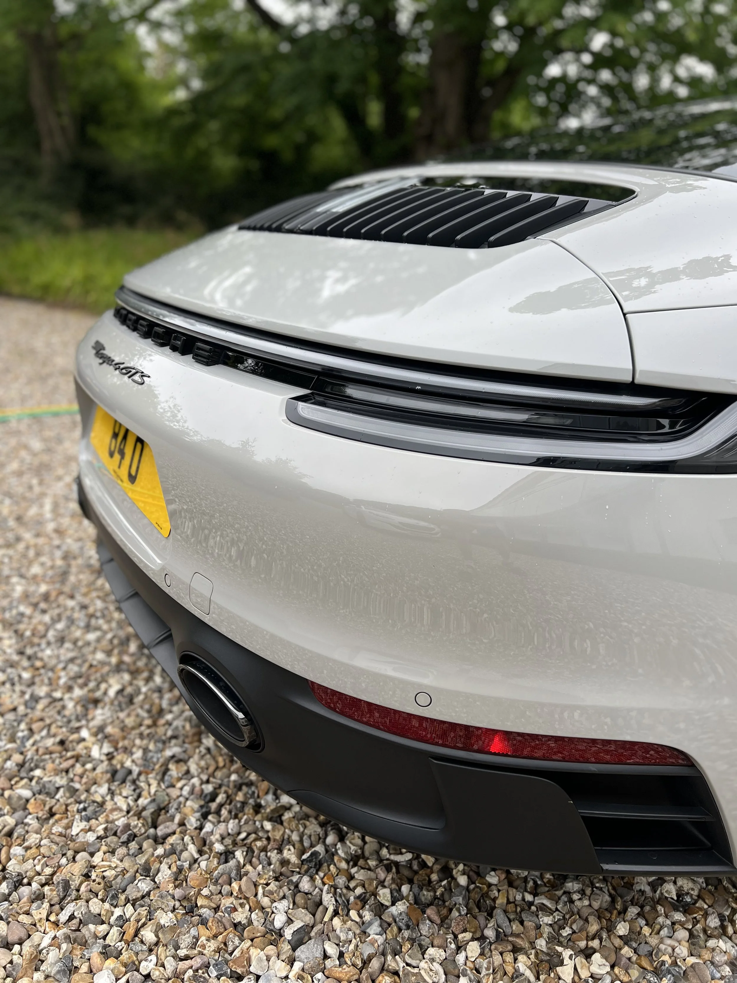 Close-up of the front right side of a white Porsche 911 Carrera S, showing the headlight, black grille vents, and part of the engine cover, parked on a gravel driveway with green trees in the background.
