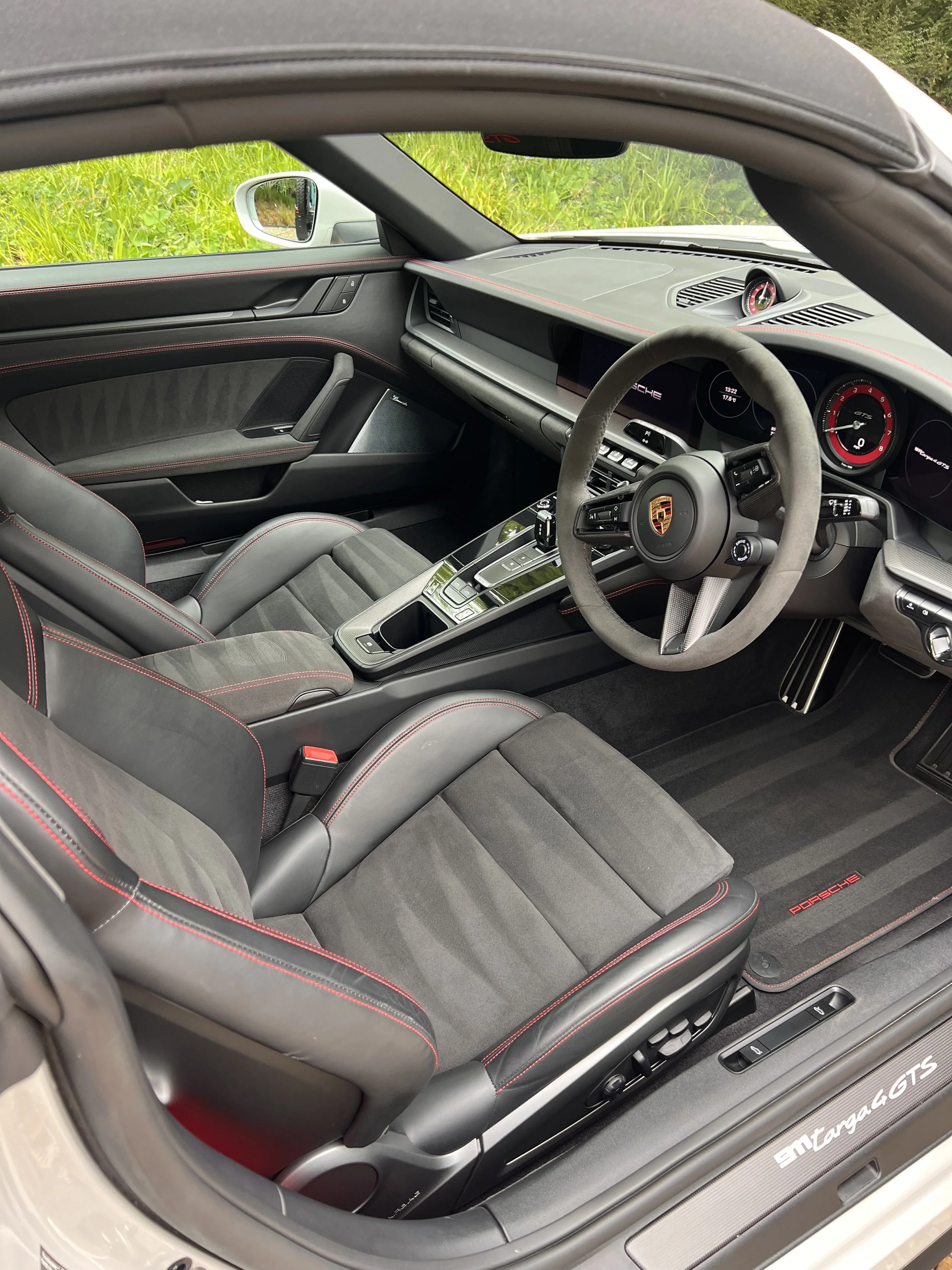 Interior of a Porsche 911 Targa 4 GTS with black seats and red contrast stitching, steering wheel with Porsche emblem, digital dashboard, and dashboard clock, view through open roof showing green grass outside.