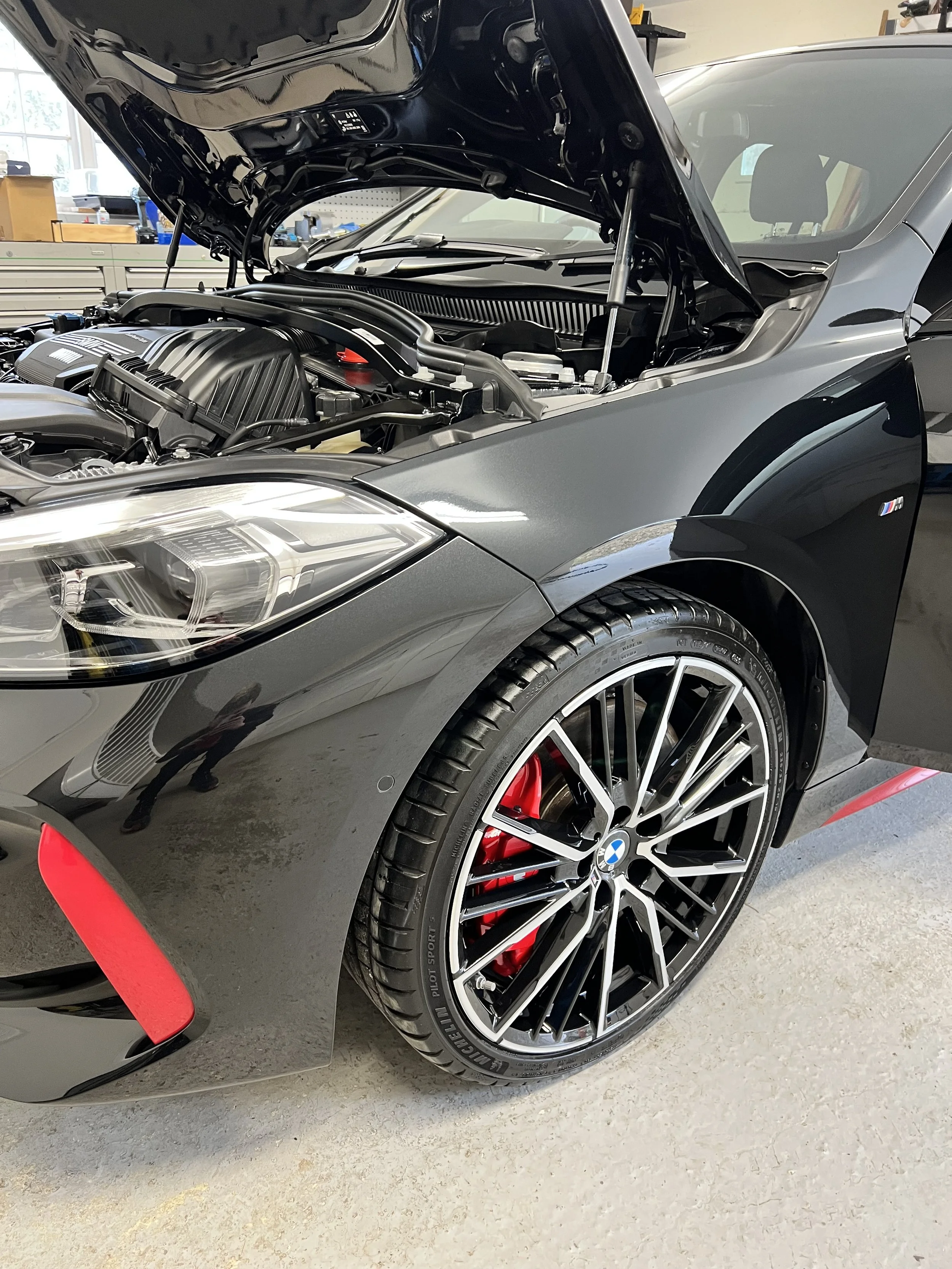 A black BMW M series sports car with a red accents around the front bumper and red brake calipers, parked inside a garage with the hood open showing the engine.