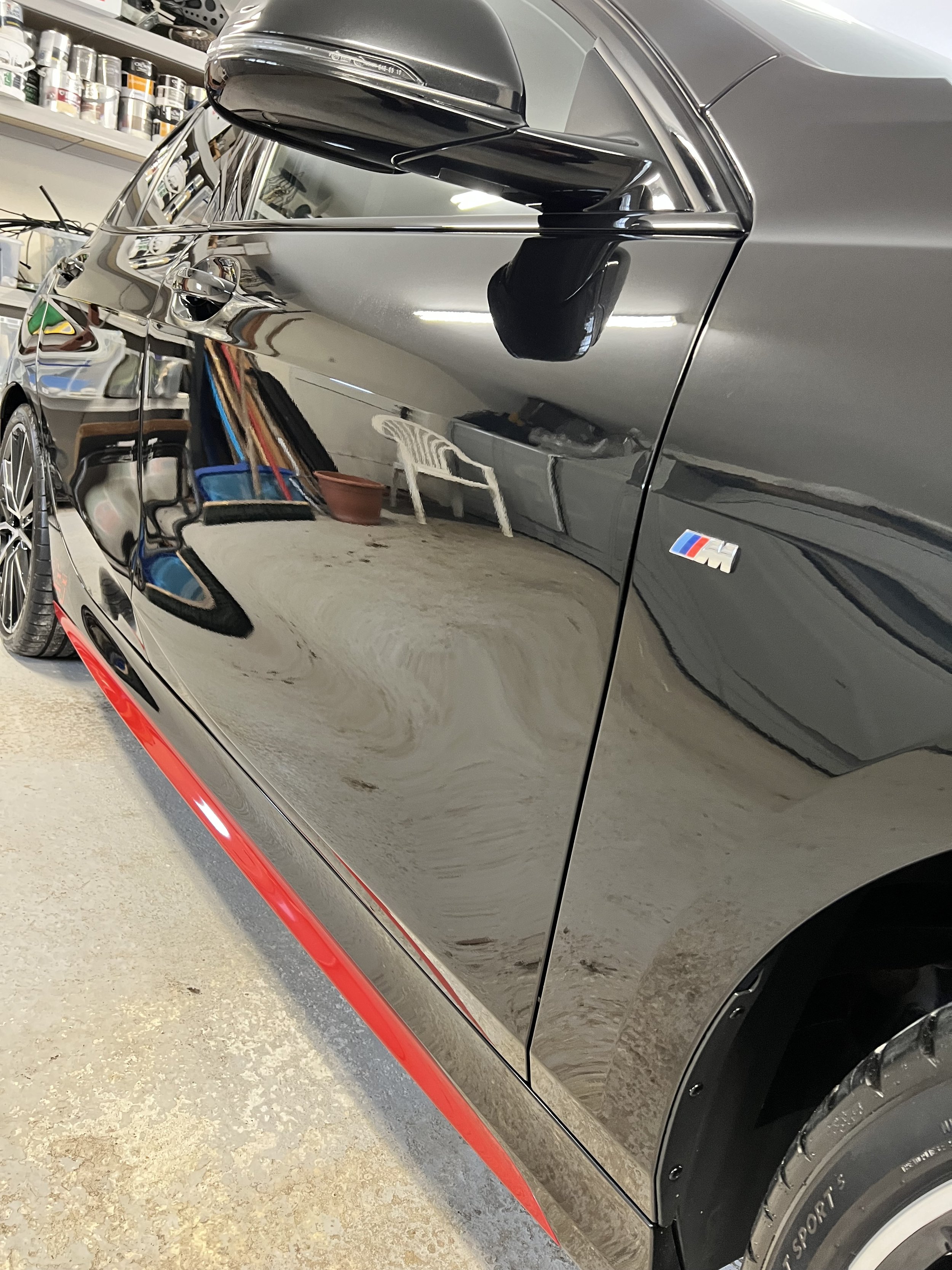 Close-up of a black BMW M Series car parked in a garage, showing the side profile with a focus on the black side mirror, and the BMW M badge on the door. The garage has shelves with paint cans and various tools reflected on the car's shiny surface.