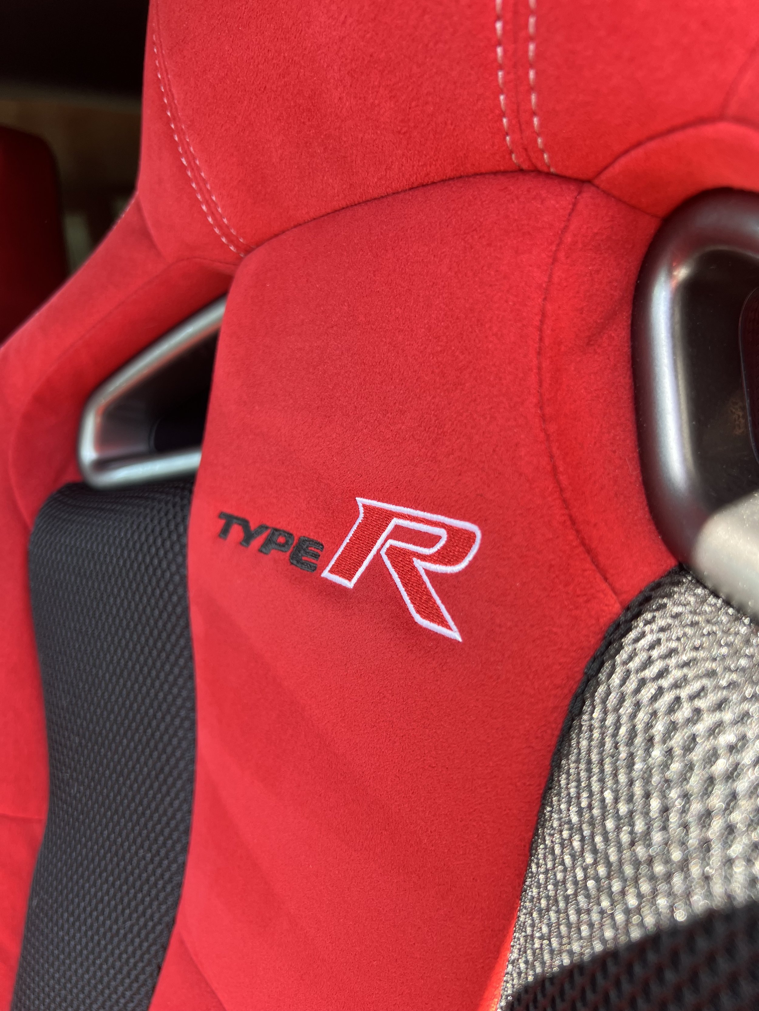 Close-up of a red racing car seat with white stitching, highlighting the "Type R" logo and black and gray fabric on the side.