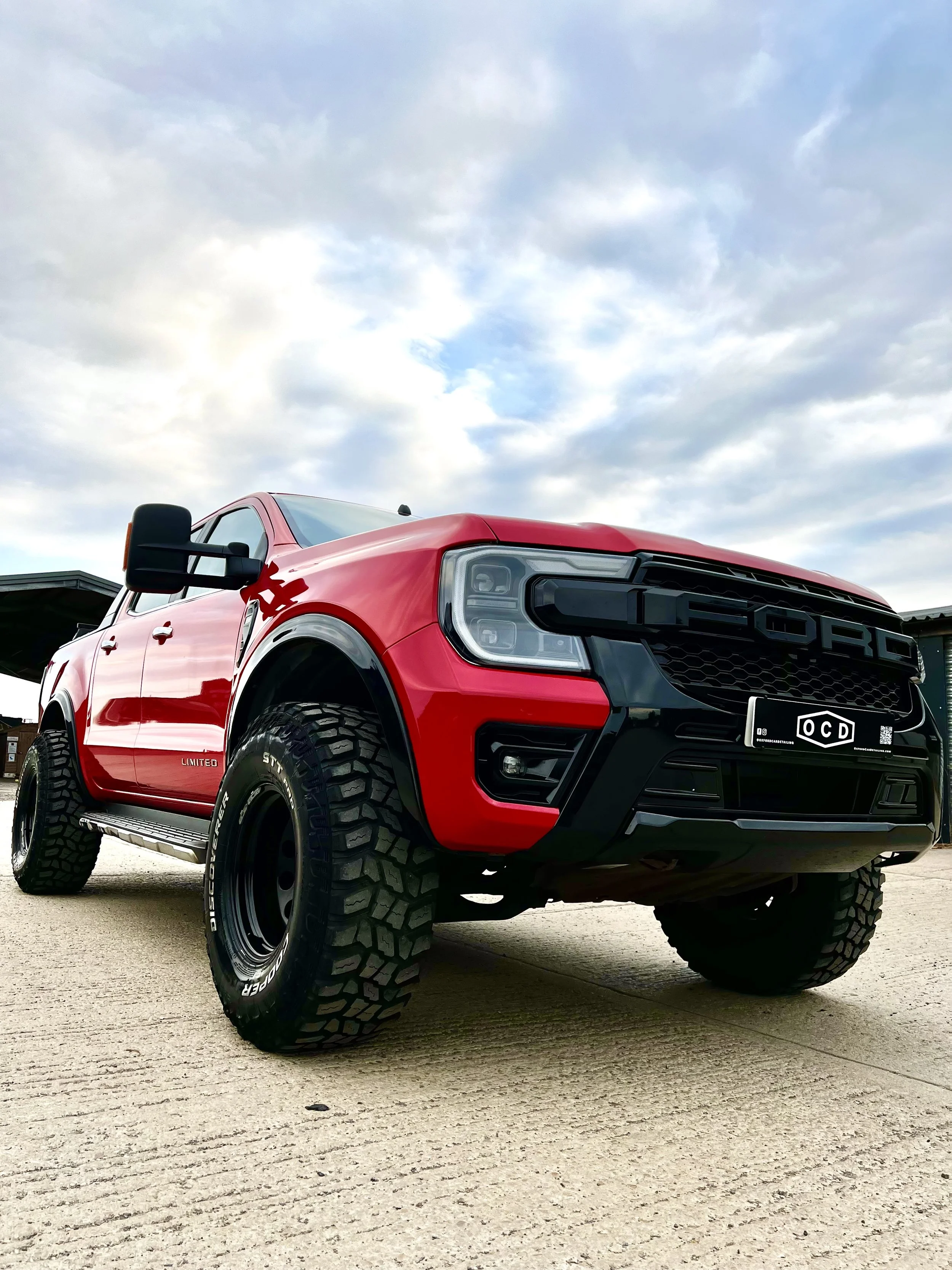 Red Ford F-150 Raptor pickup truck with off-road tires parked on concrete with cloudy sky in background.
