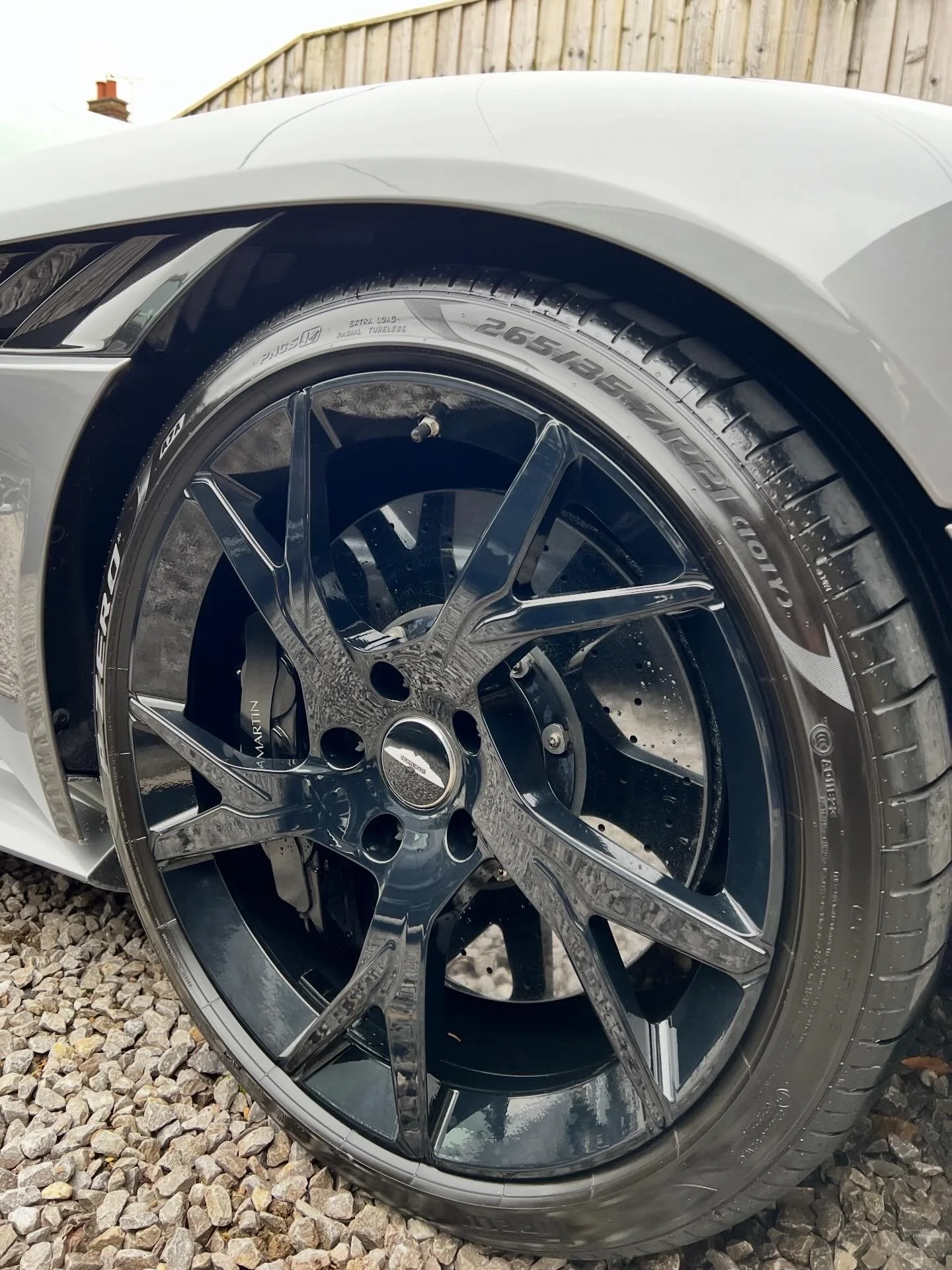 Close-up of a car wheel with a black rim and low-profile tire, parked on gravel, with part of a white car body and a wooden fence in the background.