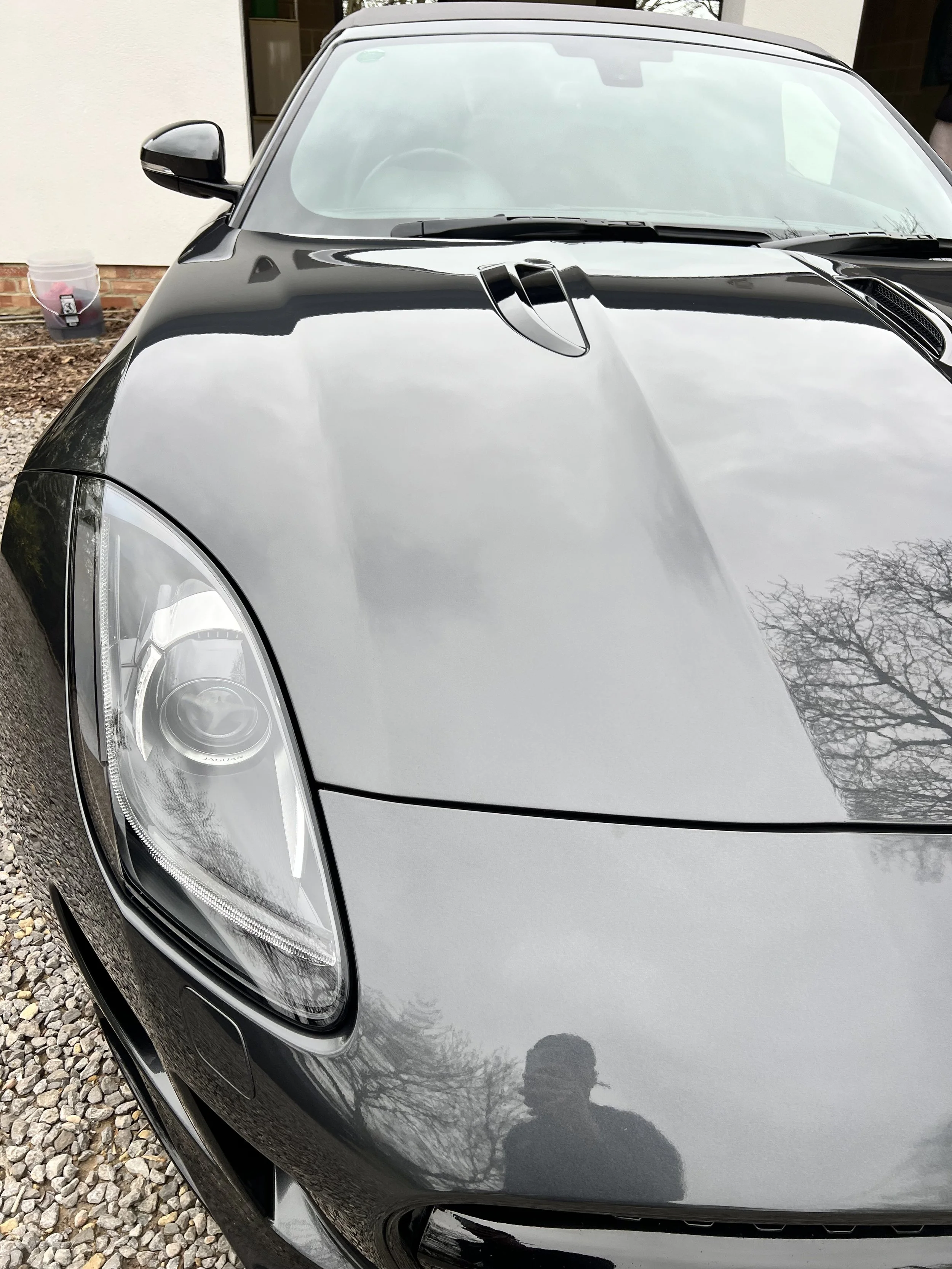 Close-up of the front part of a silver sports car, showing the headlight, hood with vents, and side mirror, parked on gravel in front of a building.