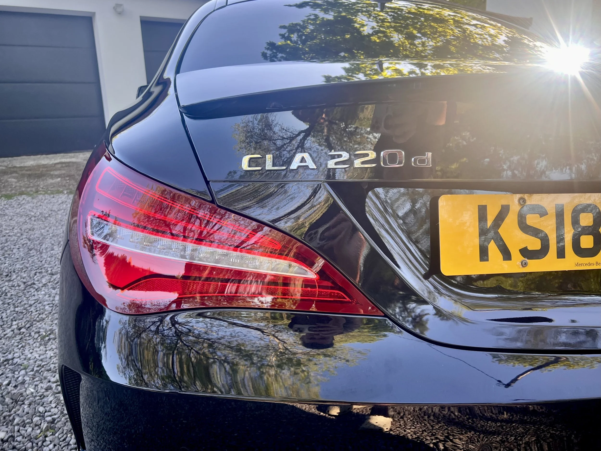 The rear end of a shiny black Mercedes-Benz CLA 220d car parked outdoors, showing the tail light, model badge, and part of the license plate, with reflected trees and sunlight on its surface.