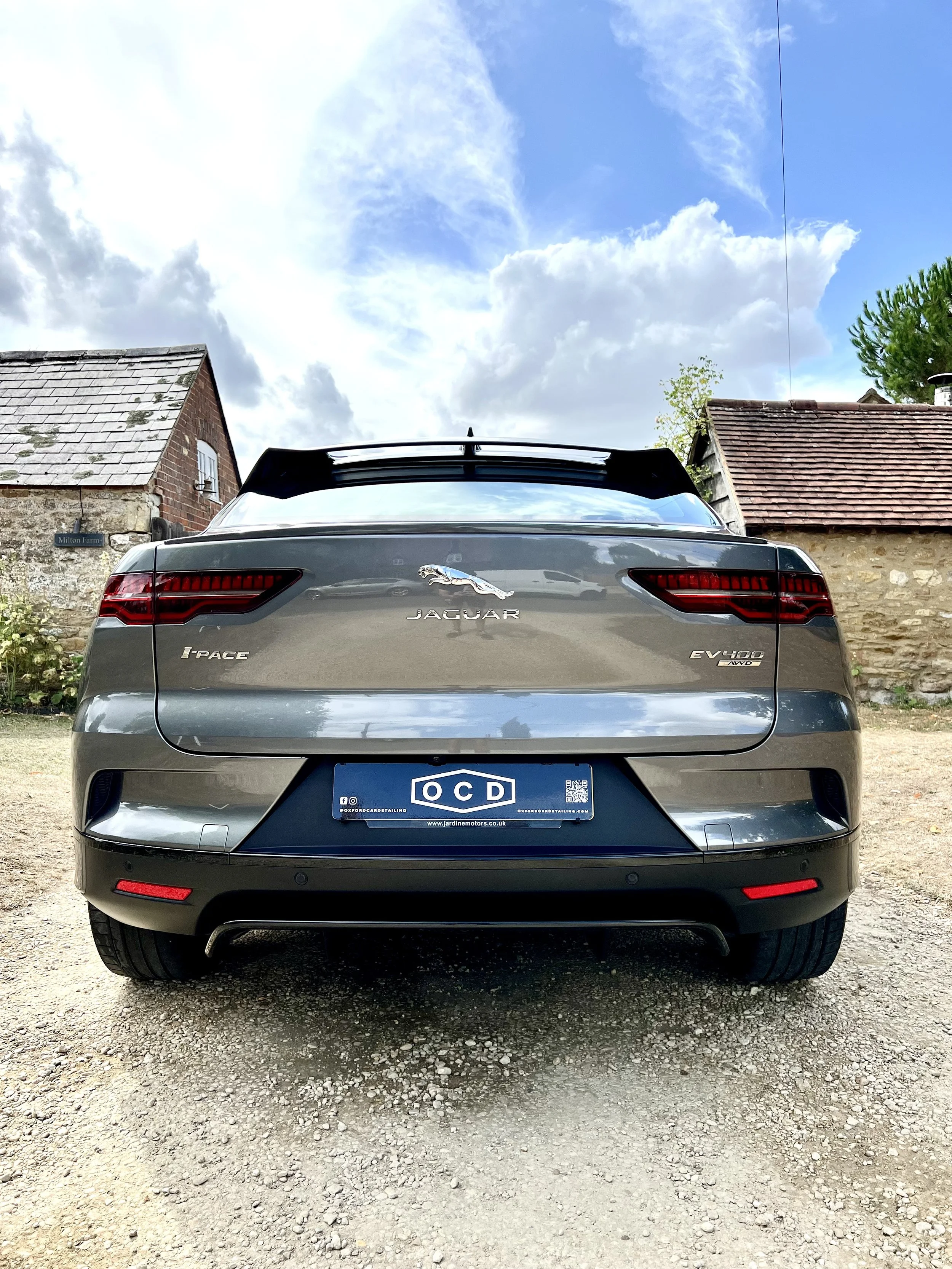 Rear view of a gray Jaguar I-PACE electric vehicle parked on a gravel surface, with a barn and a house in the background and a cloudy sky above.