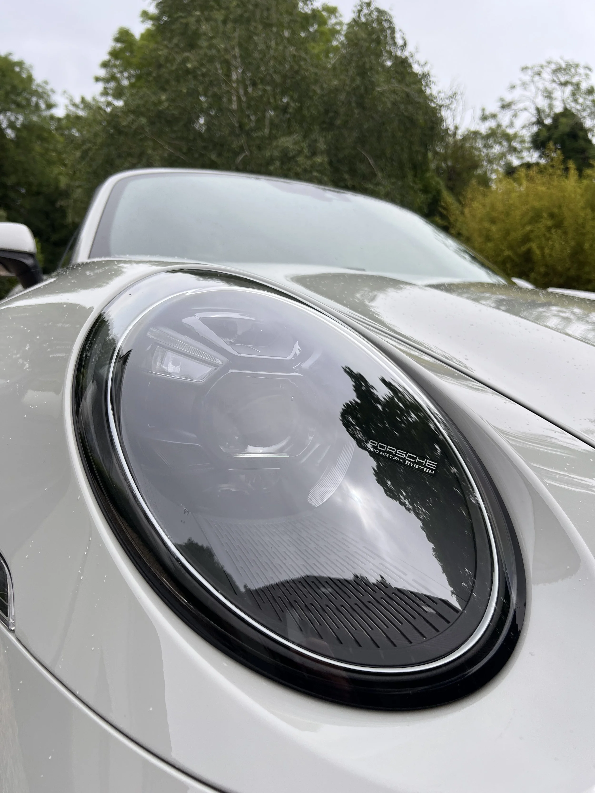 Close-up of a silver Porsche car hood with its headlight and windshield visible, reflecting surrounding trees and cloudy sky.