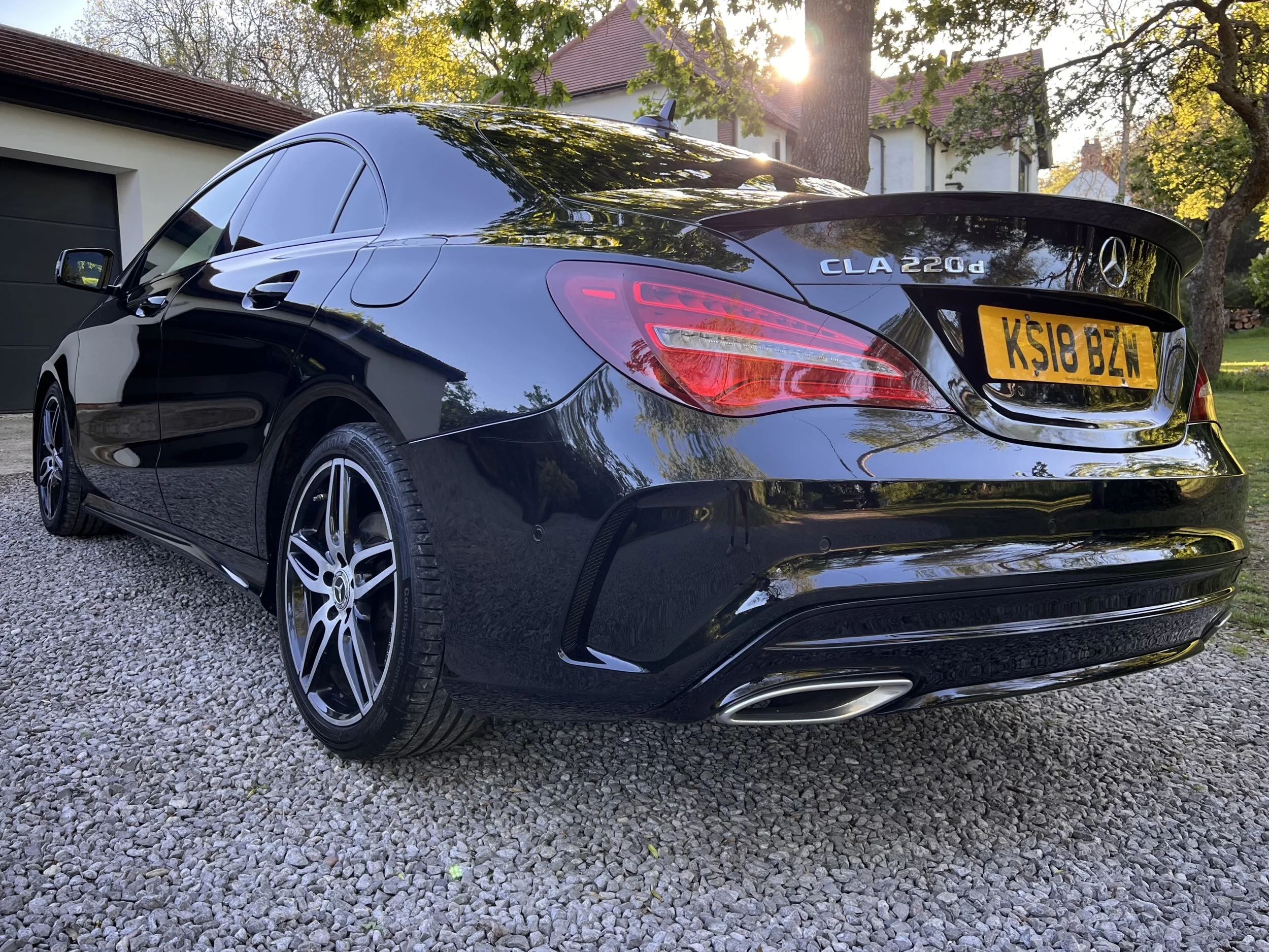 A black Mercedes-Benz CLA 220d parked on a gravel driveway in front of a house with trees and a garage. The car’s rear and passenger side are visible, with sunlight shining through the trees.