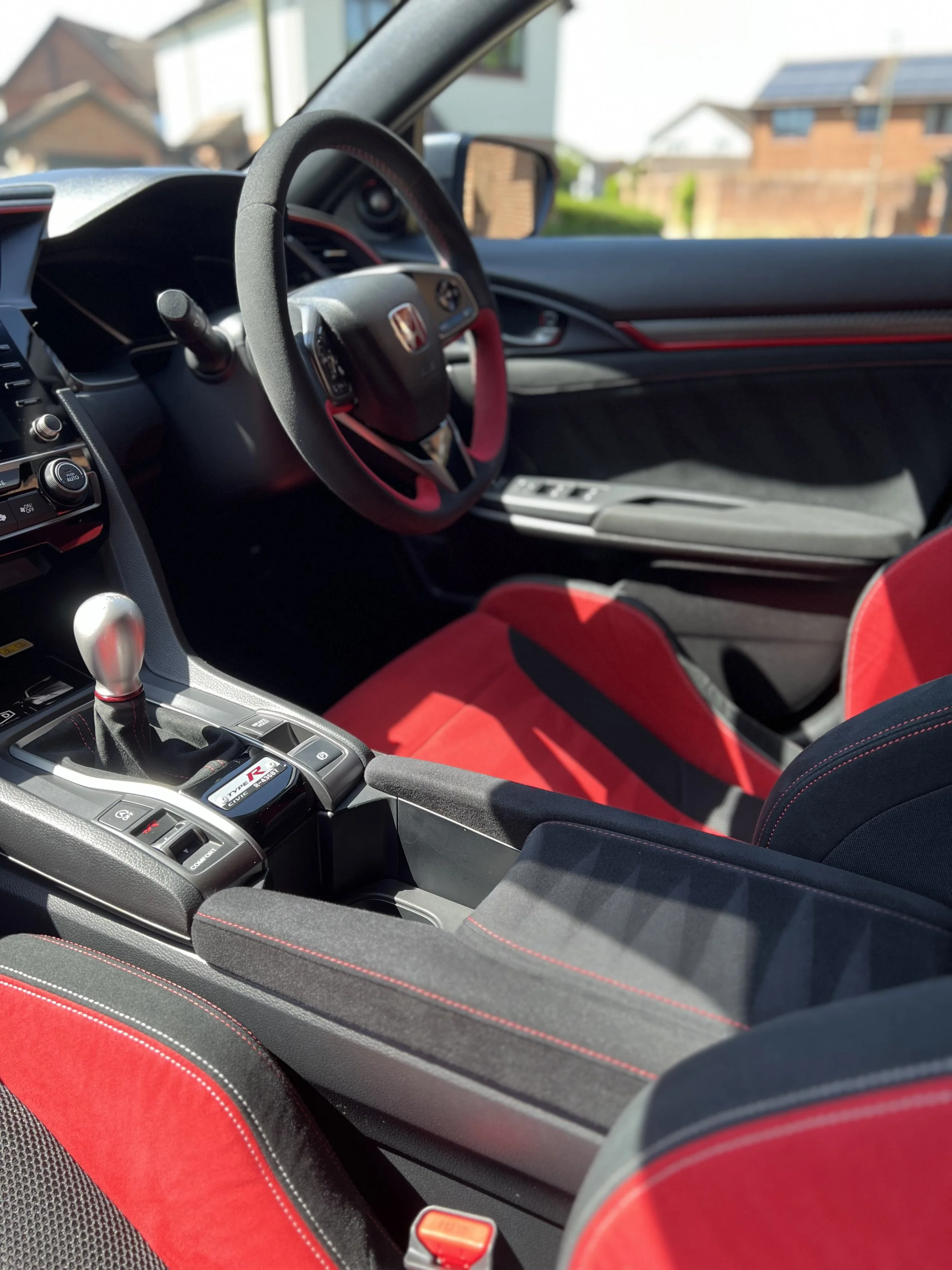 Inside view of a car's interior showing a racing steering wheel with red and black accents, a gear shift, and red seat upholstery.