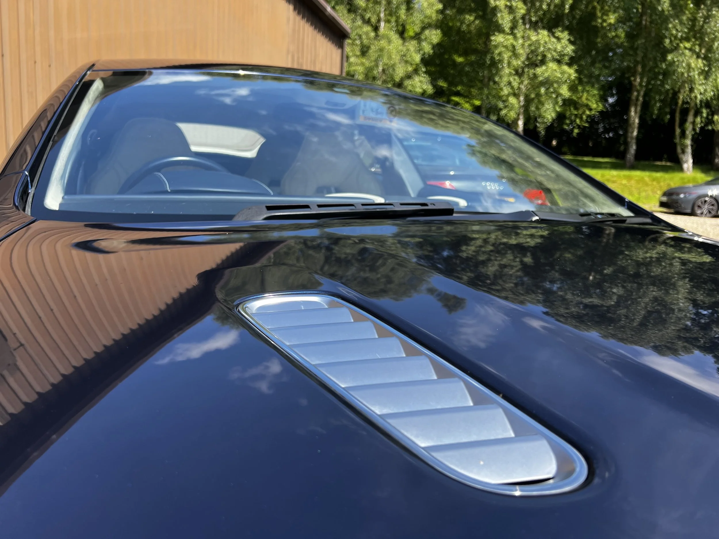 A close-up of the hood of a black sports car showing metallic vents, with a view of the windshield and interior, reflecting the sky and trees, and a wooden building in the background.