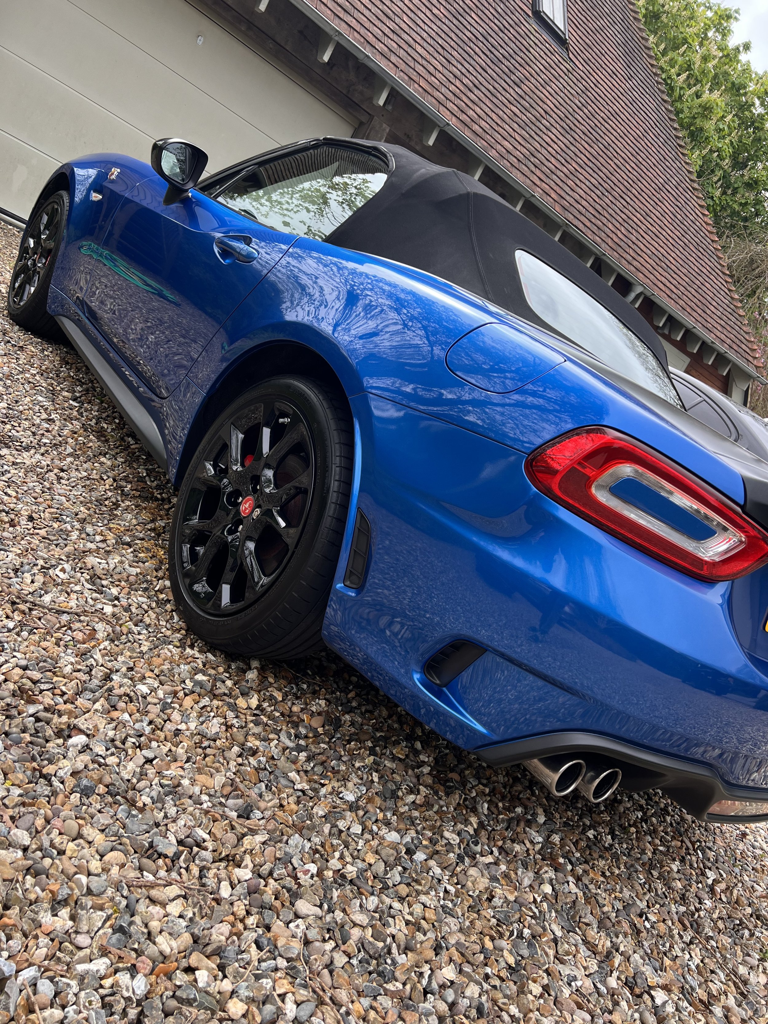 A blue convertible sports car parked on a gravel driveway next to a building with a brick wall and a gray garage door.