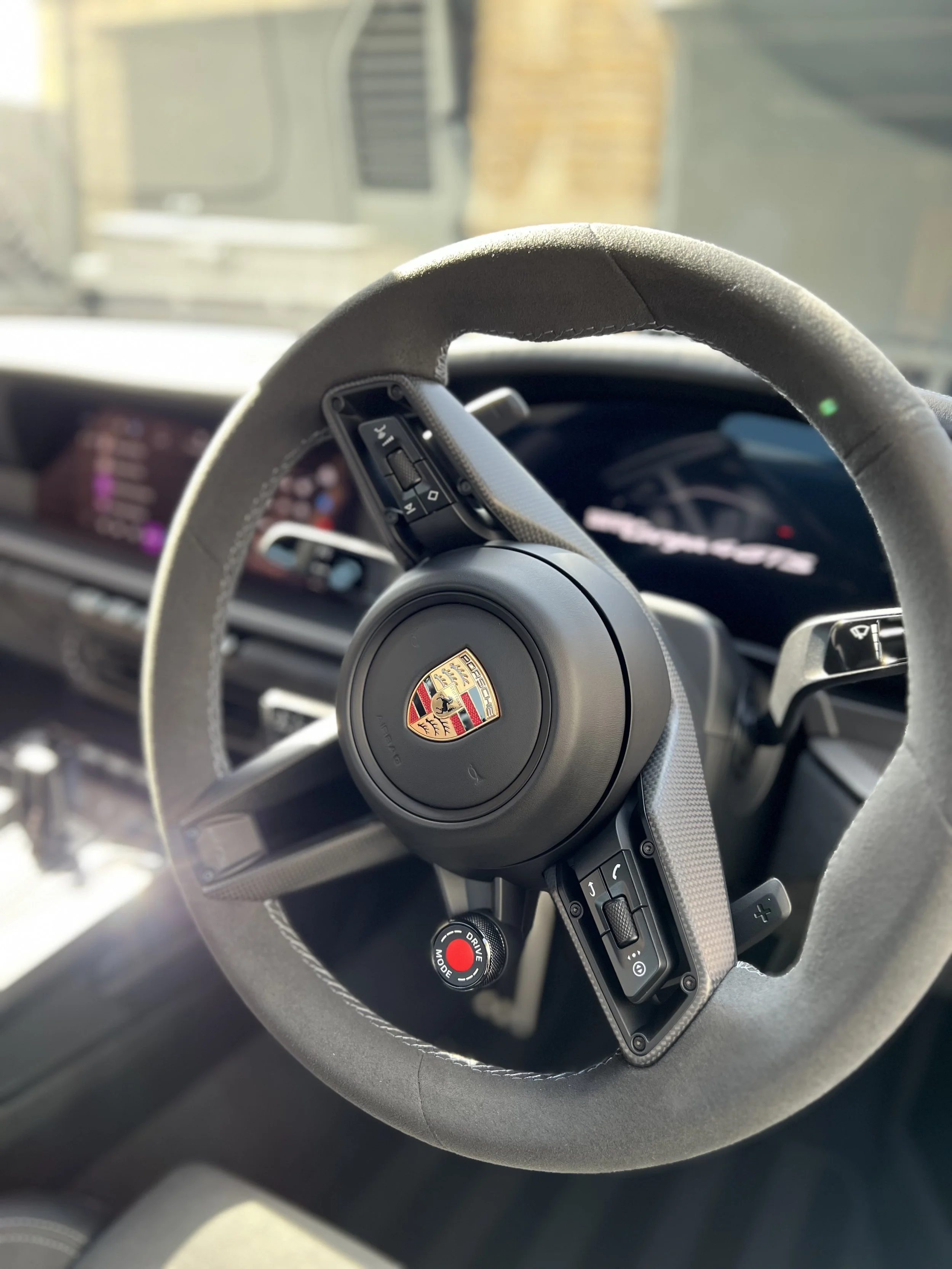 Close-up of a Porsche steering wheel with a Porsche logo in the center, and various control buttons on the left side, in a luxury car interior.