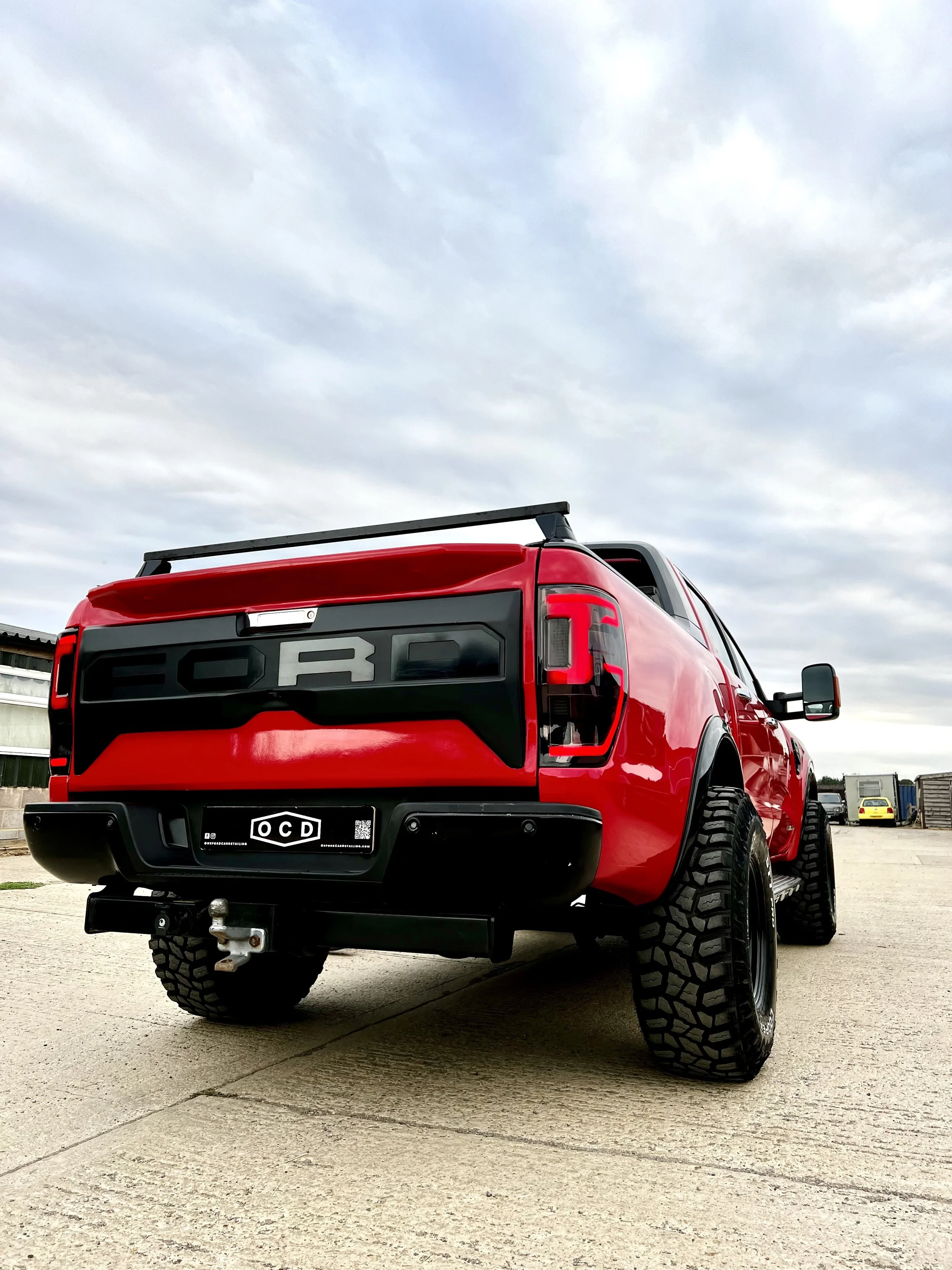 Red Ford truck with black tailgate and large tires parked on a concrete lot under cloudy sky.