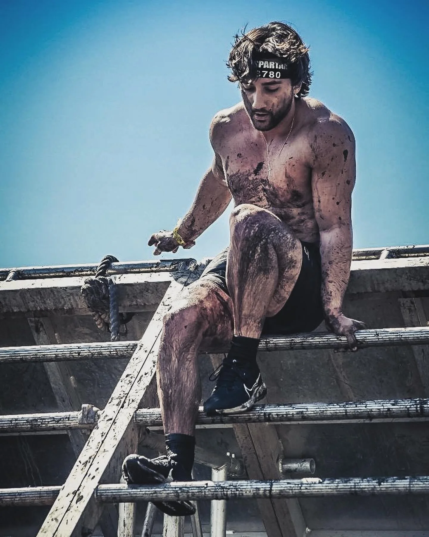 A shirtless man wearing a headband, shorts, and running shoes, climbing over a wooden obstacle during a mud race or obstacle course event, with mud on his body and a clear blue sky in the background.