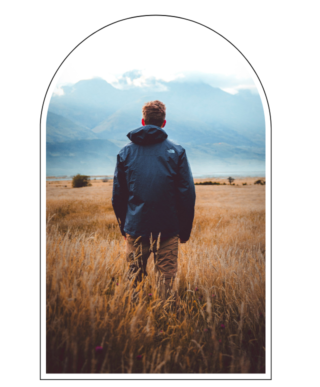 A person in a dark jacket and brown pants standing in a golden grassy field, looking at mountains and a cloudy sky.