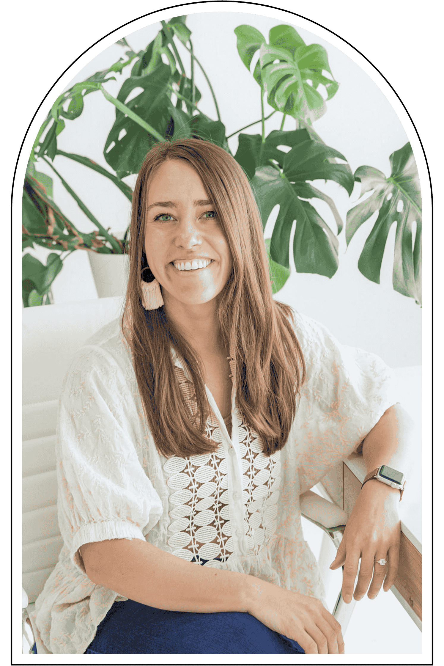 Female trauma therapist with long brown hair smiling, wearing a white embroidered blouse, earrings, and a smartwatch, sitting in front of green houseplants.
