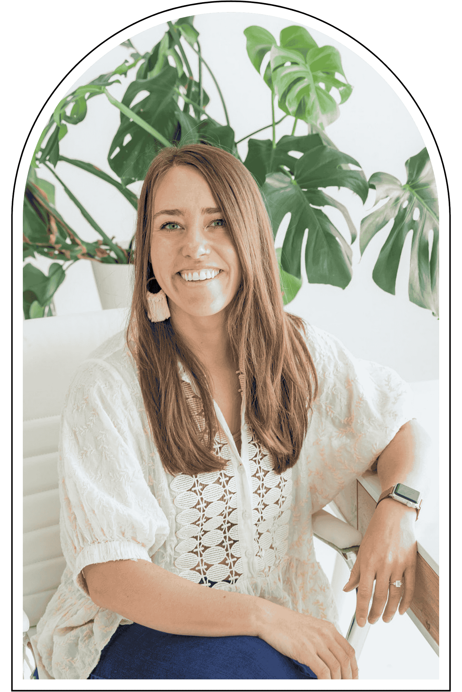 A woman with brown hair, smiling, wearing a white embroidered blouse, sitting in front of large indoor green plants, with a smartwatch on her left wrist and a ring on her right hand.