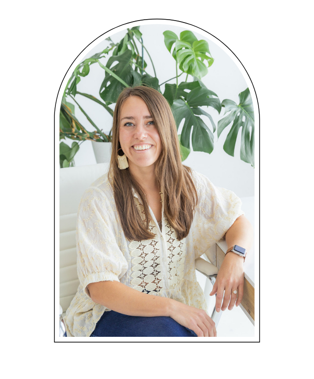 A smiling woman with light brown hair, wearing a cream-colored embroidered blouse and a smartwatch, seated at a desk with large green houseplants in the background.