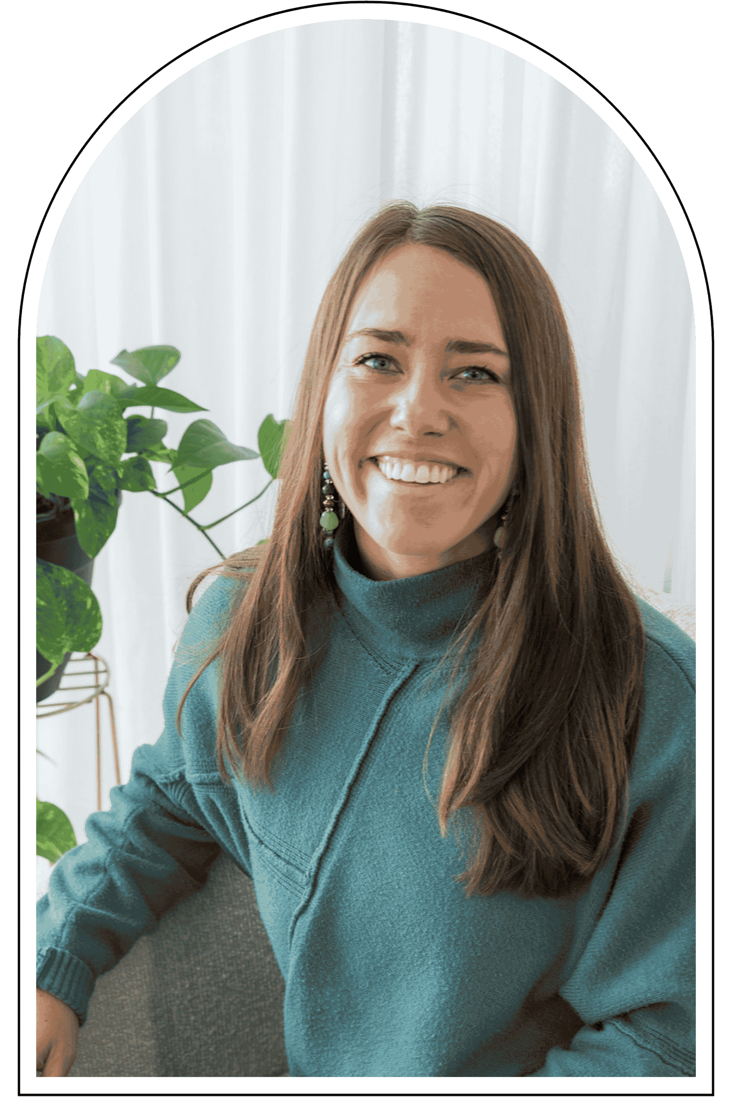 A woman with long brown hair, wearing a teal sweater, and earrings, smiling at the camera in a well-lit indoor setting with green plants in the background.