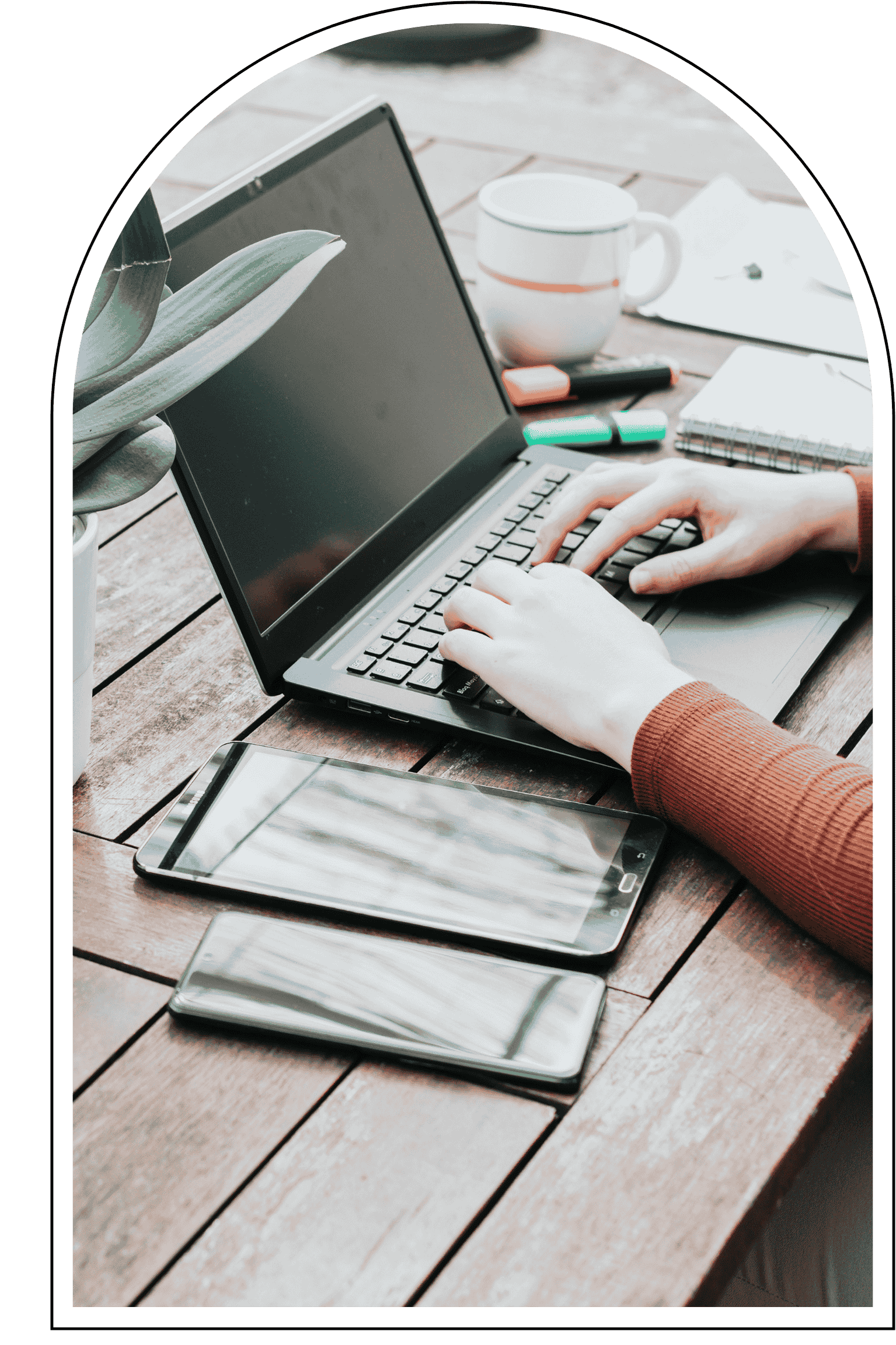 Hands typing on a laptop keyboard on a wooden table, with a smartphone, tablet, coffee mug, notebooks, and highlighters nearby.