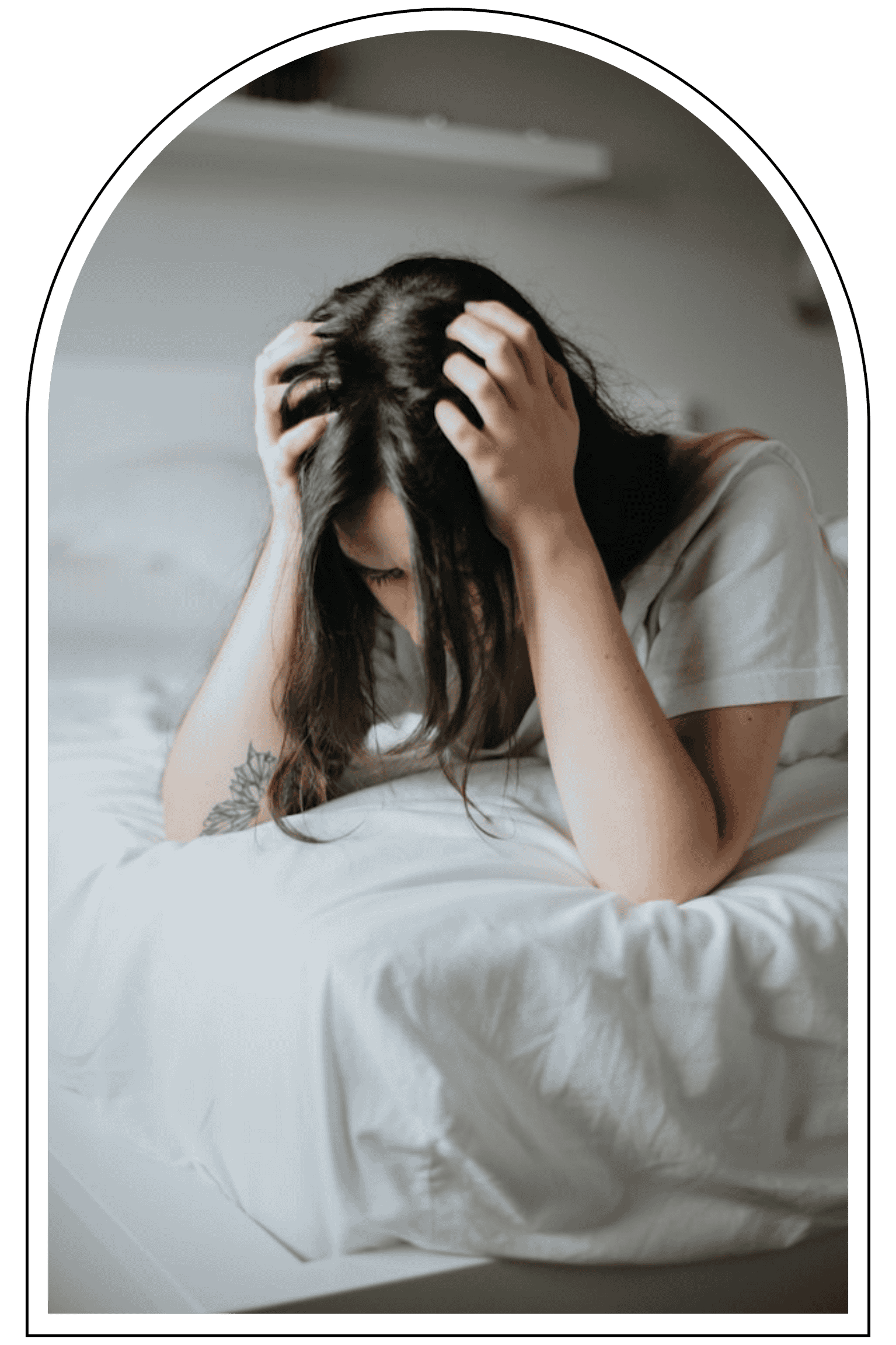 A woman with dark hair and a tattoo on her arm, sitting on a bed with white sheets, holding her head with both hands, appearing distressed or upset.