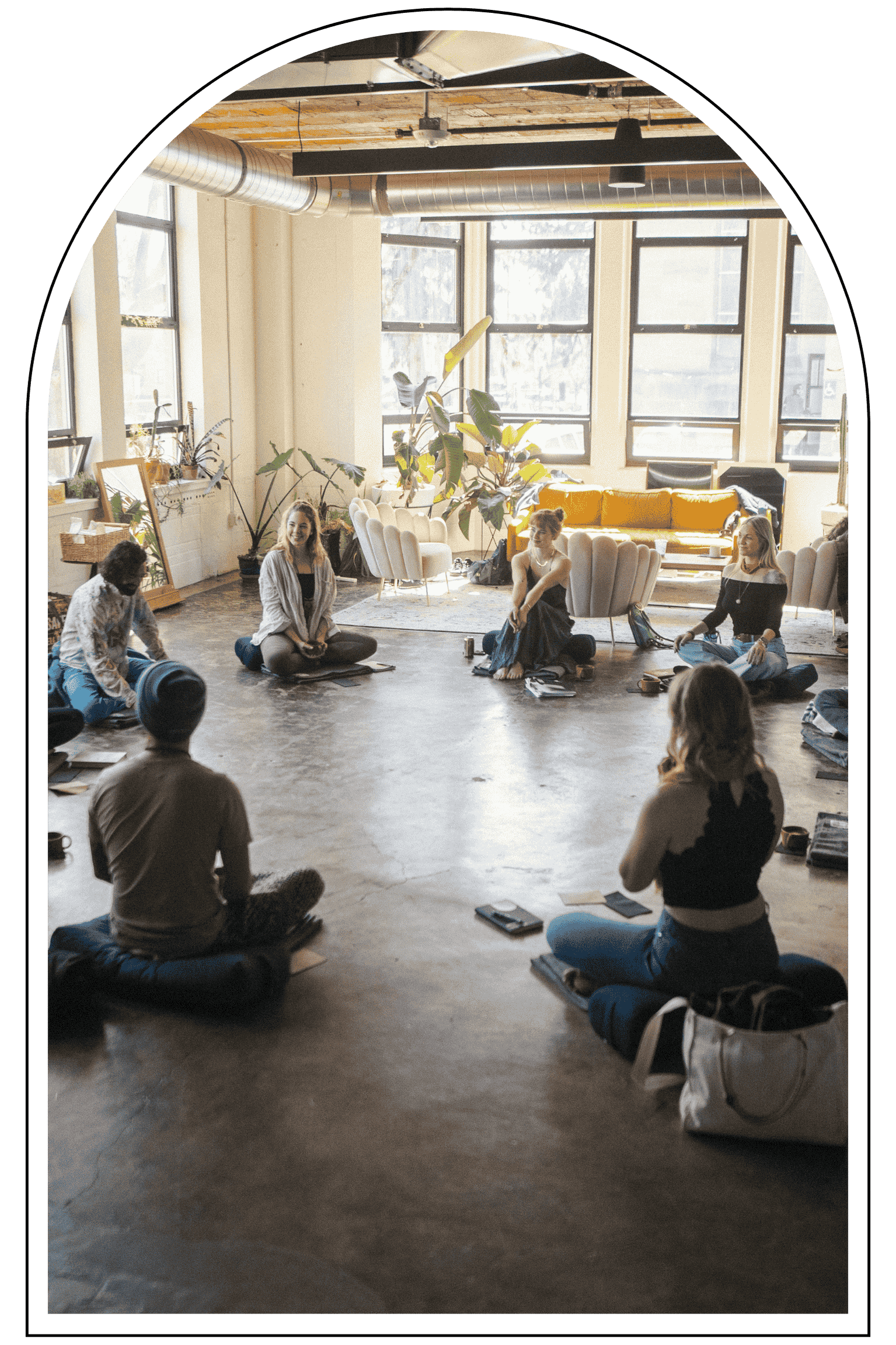 Group of people sitting on the floor in a spacious, well-lit room with large windows, engaged in a discussion or class.