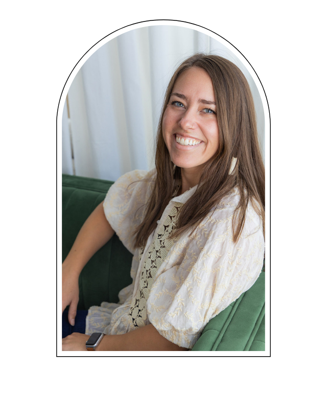 A smiling woman with long brown hair sitting on a green couch near white curtains.